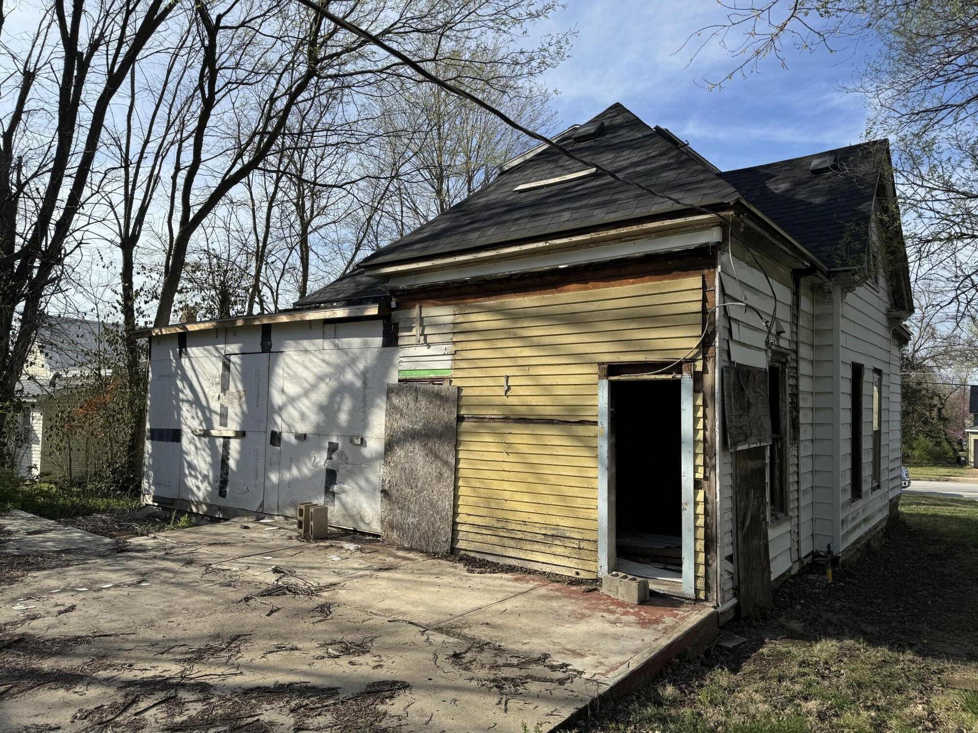 Weathered house with damaged siding, boarded windows, and an open doorway beside a concrete patio.