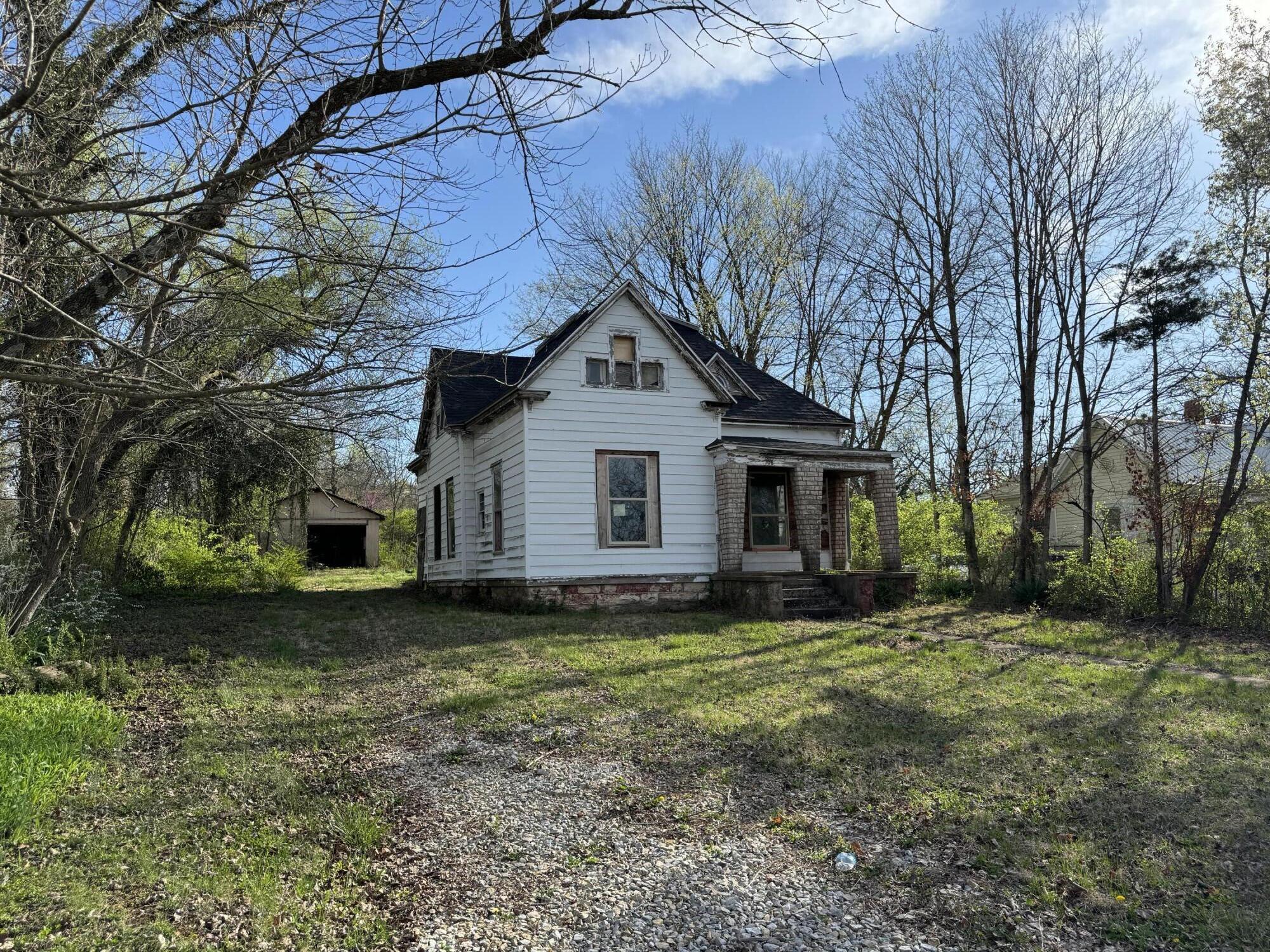 Older white-sided house with a steep gabled roof, brick porch supports, and a gravel driveway.