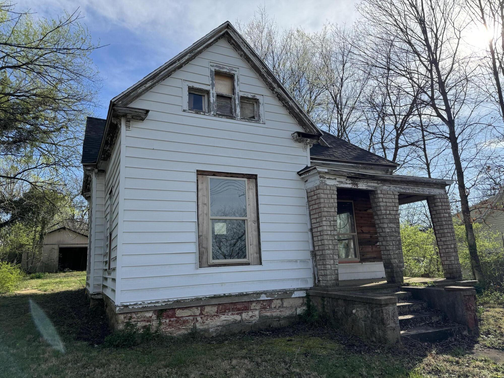 Weathered white two-story house with a steep gable and stone-column porch, surrounded by bare trees.