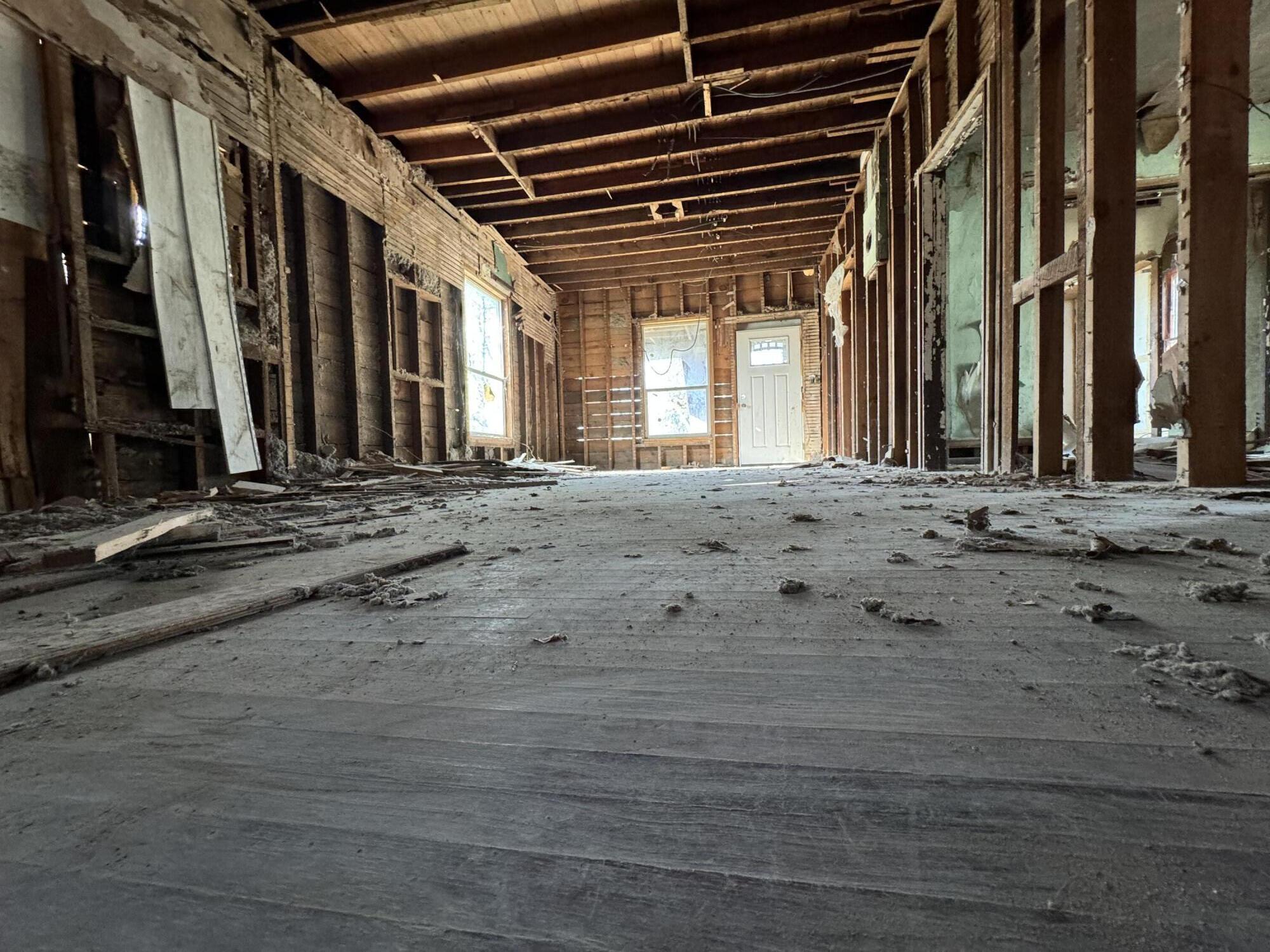 Stripped interior with exposed wooden framing, debris-strewn floor, and a door and windows at the far end.