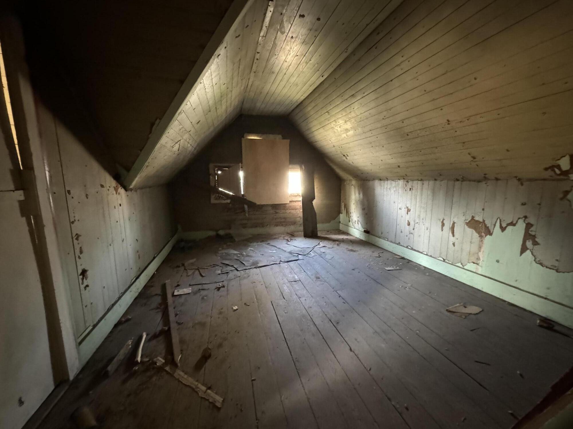 Empty attic with a steep wood-plank ceiling, peeling paneling, and debris on the floor.