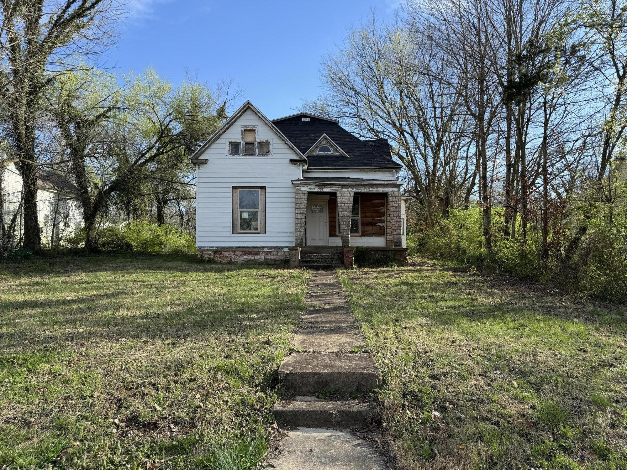 White two-story house with a dark roof and stone porch, beyond a concrete walkway and grassy yard.