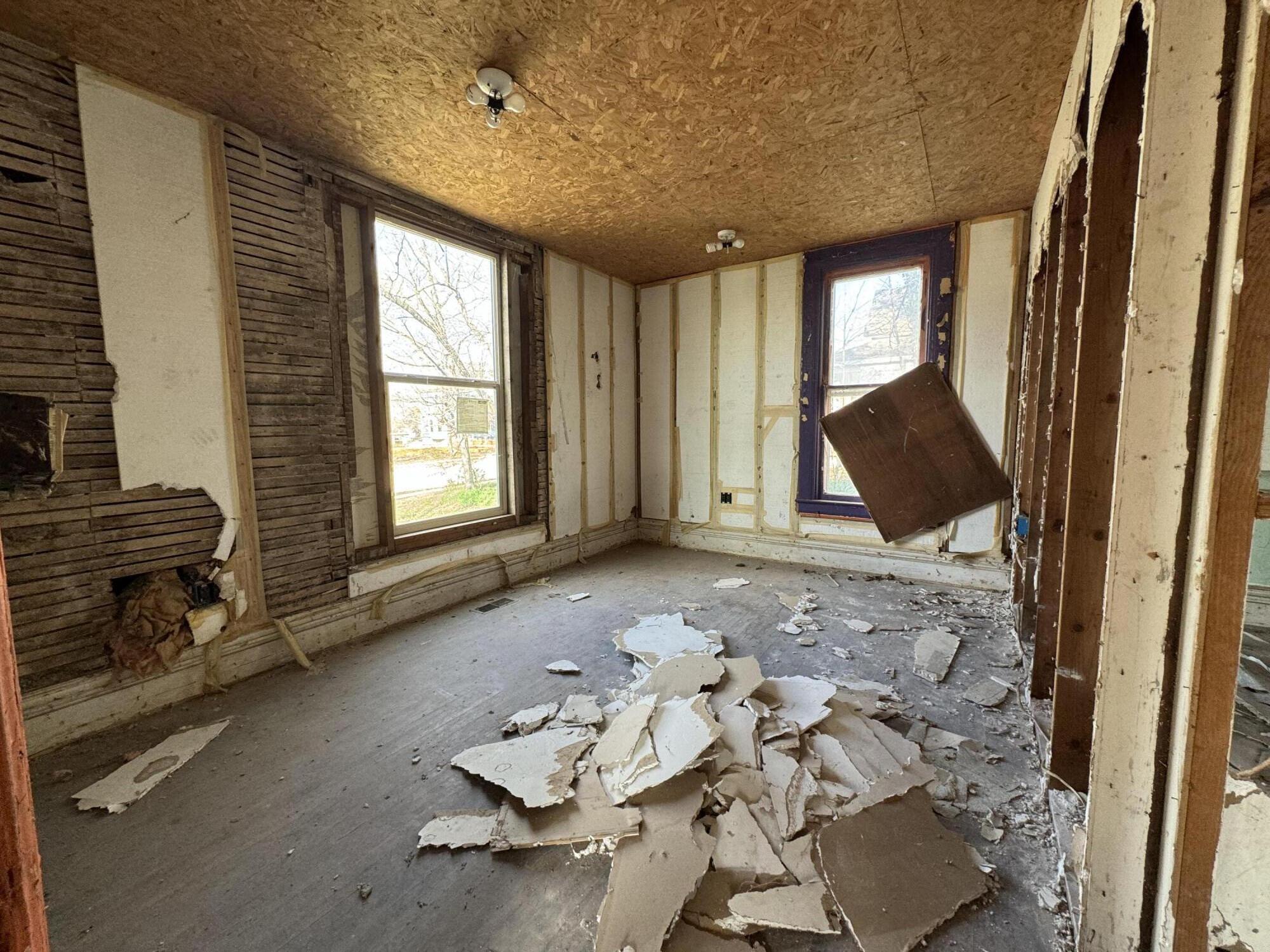 Room under renovation with exposed wall framing, two windows, and piles of drywall debris.
