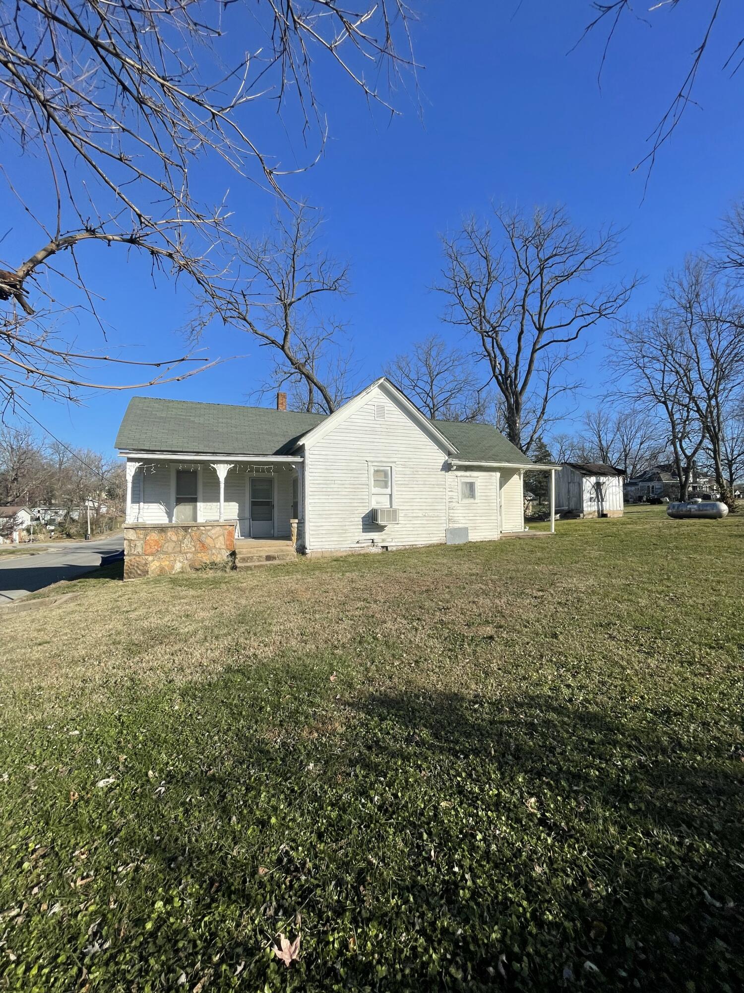 White one-story house with a covered porch, stone porch wall, and detached shed beside a grassy yard.
