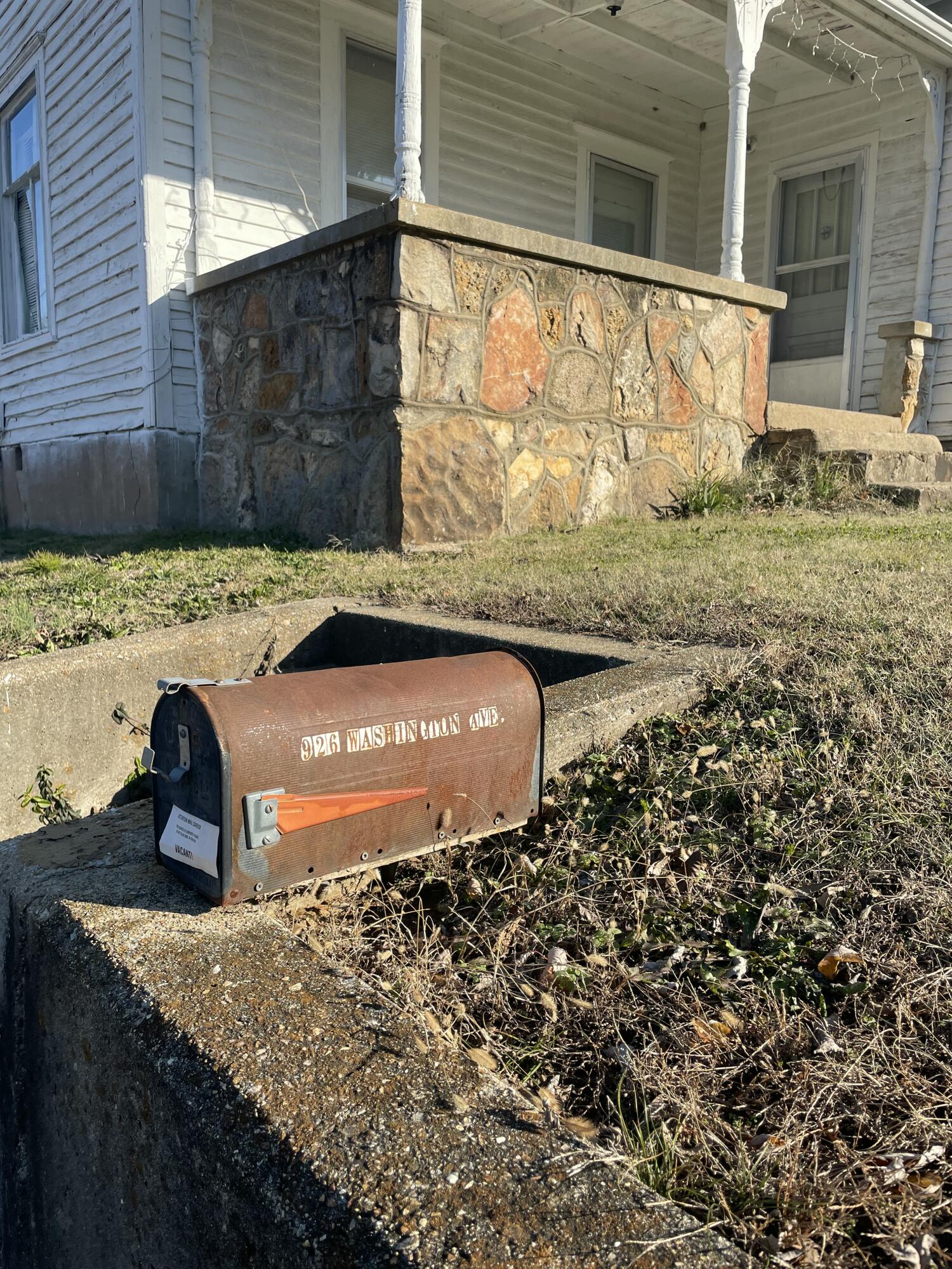 White clapboard house with a stone porch behind a rust-colored mailbox beside an overgrown walkway.