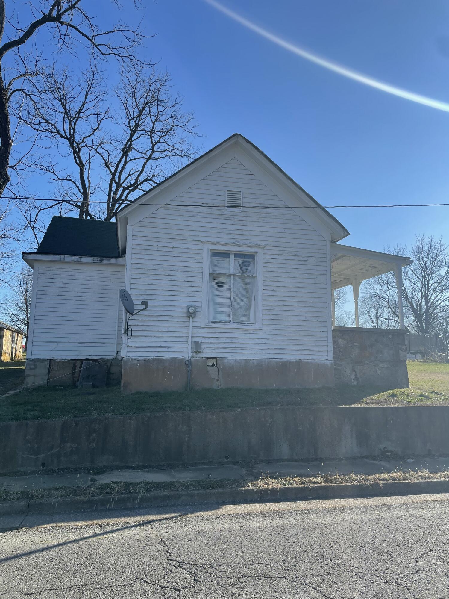 White clapboard house with a steep gable, side window, and covered porch beside a street.