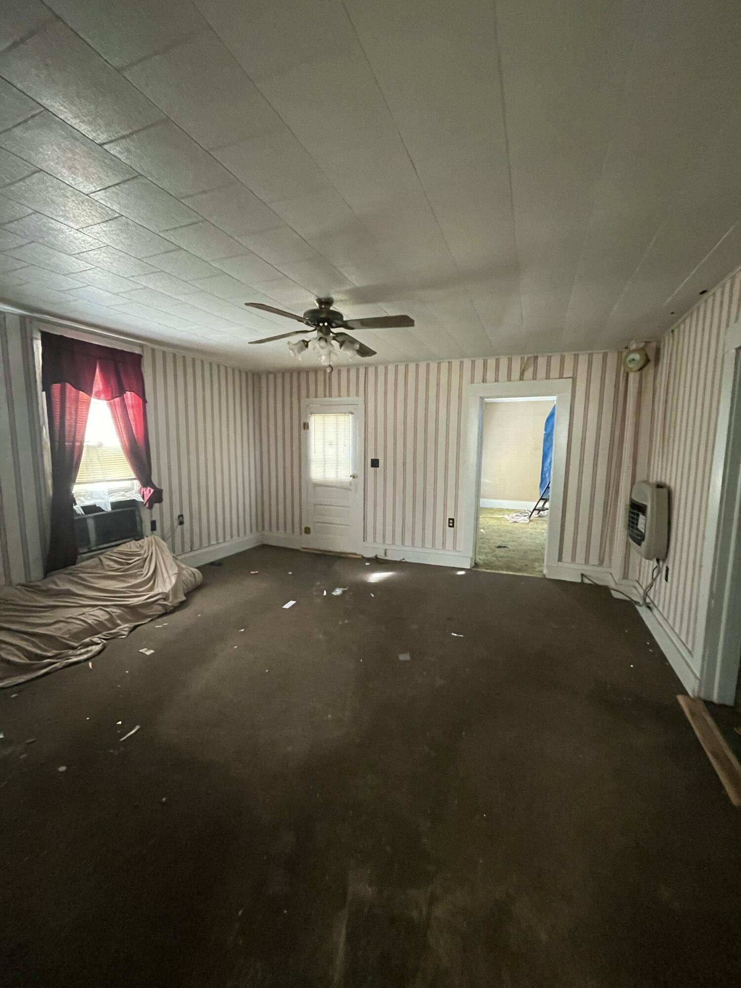 Vacant room with striped wallpaper, dark carpet, ceiling fan, and red curtains over the window.