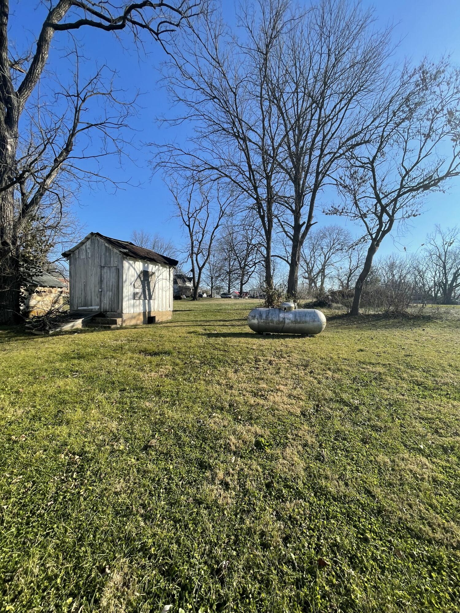 Grassy yard with a weathered wooden shed and horizontal propane tank beneath leafless trees.