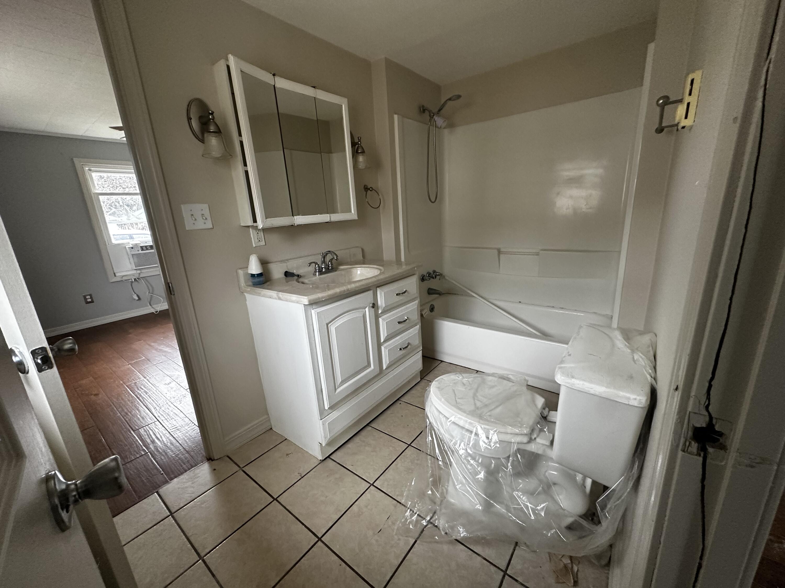 Bathroom with a white vanity, tub-shower, and toilet covered in protective plastic.
