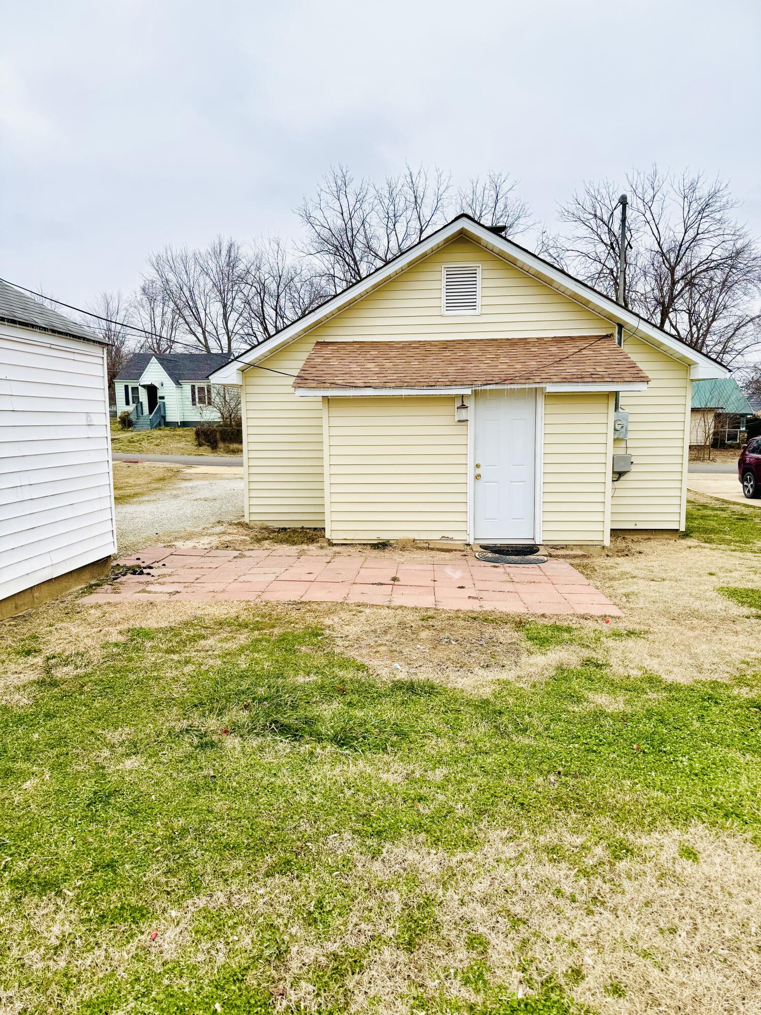 Pale yellow detached outbuilding with a white door, brown awning, and paved patio.