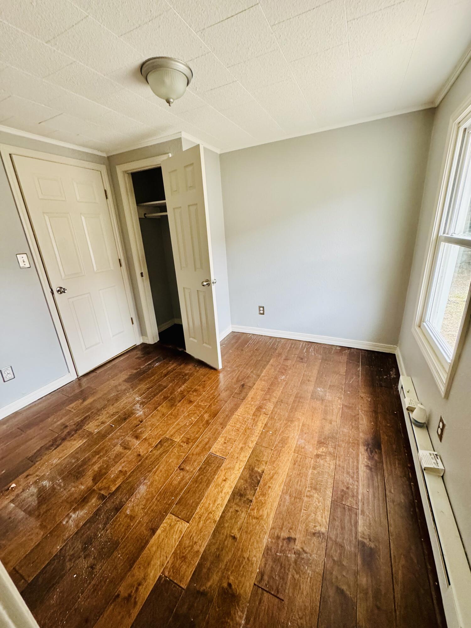 Empty bedroom with worn wood floors, an open closet, and a large window.