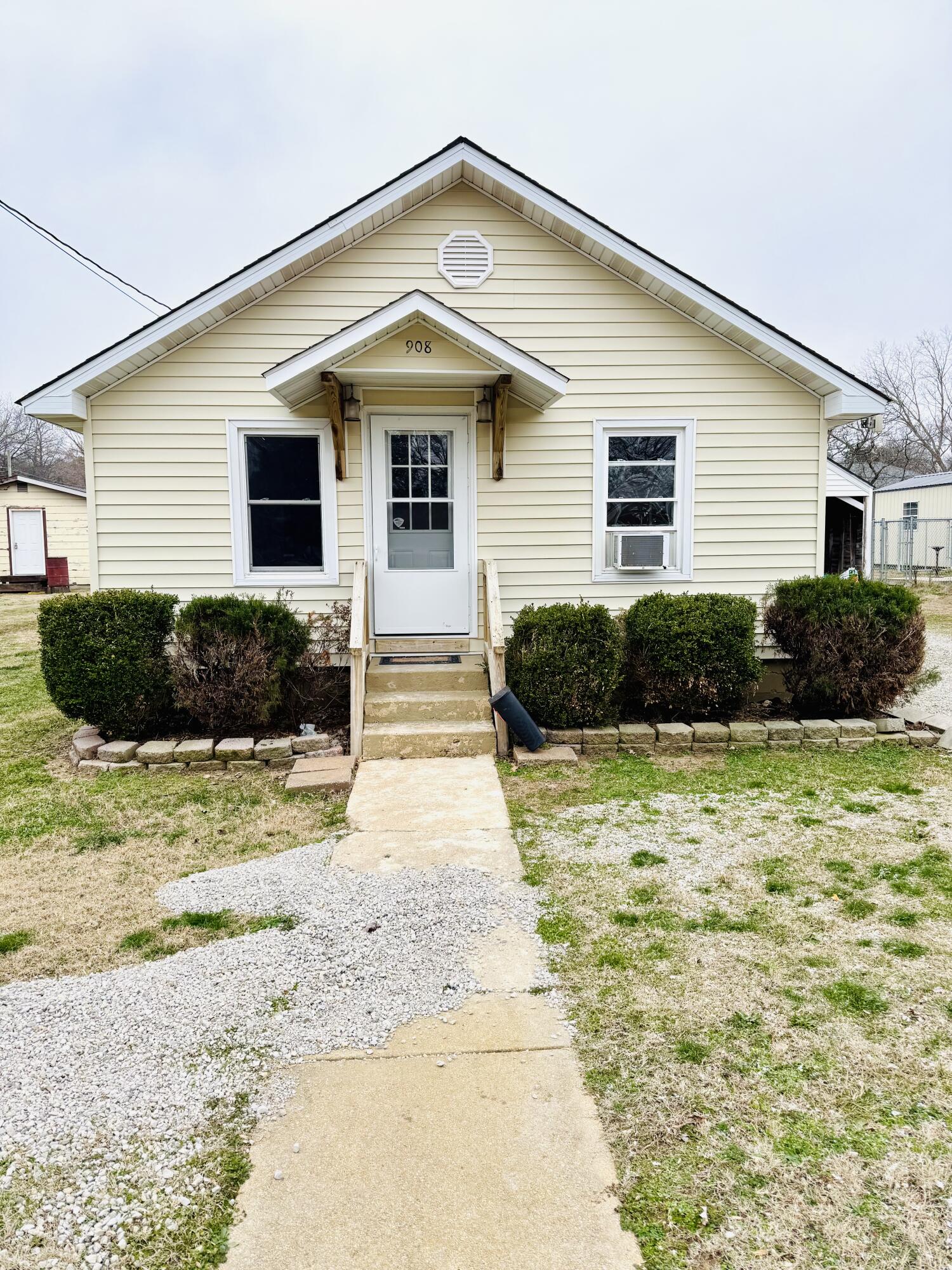 Cream-colored house with a large front gable, central entry, trimmed shrubs, and a concrete walkway.