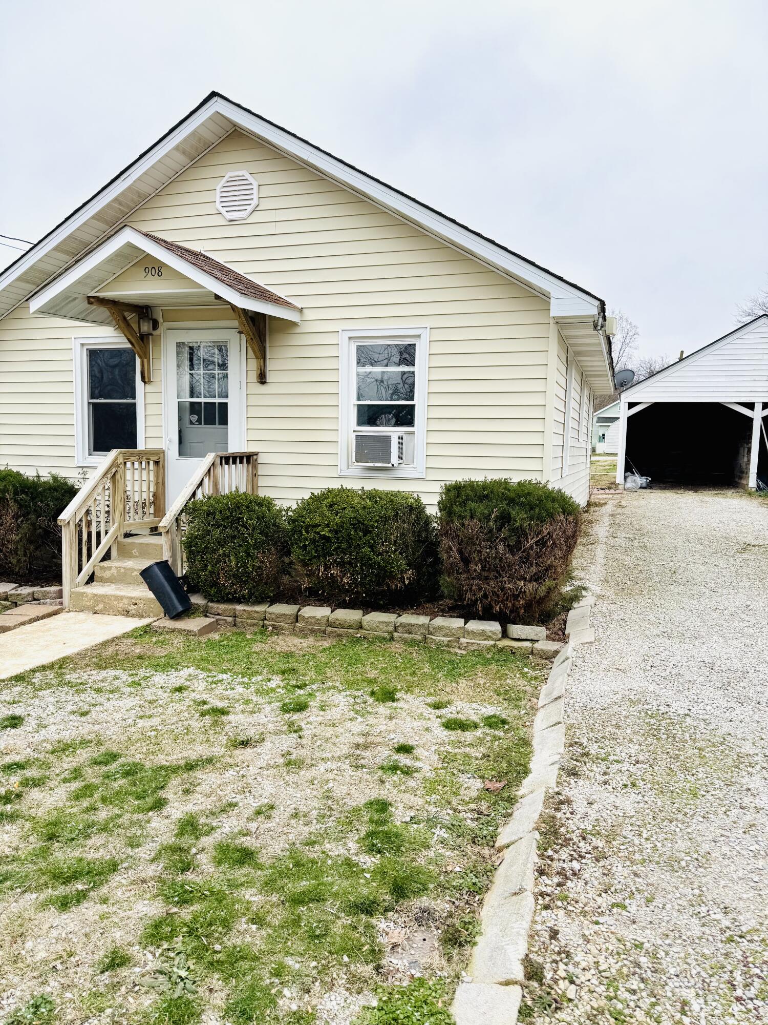 Pale-yellow house with a covered porch, front shrubs, and a gravel drive beside a carport.