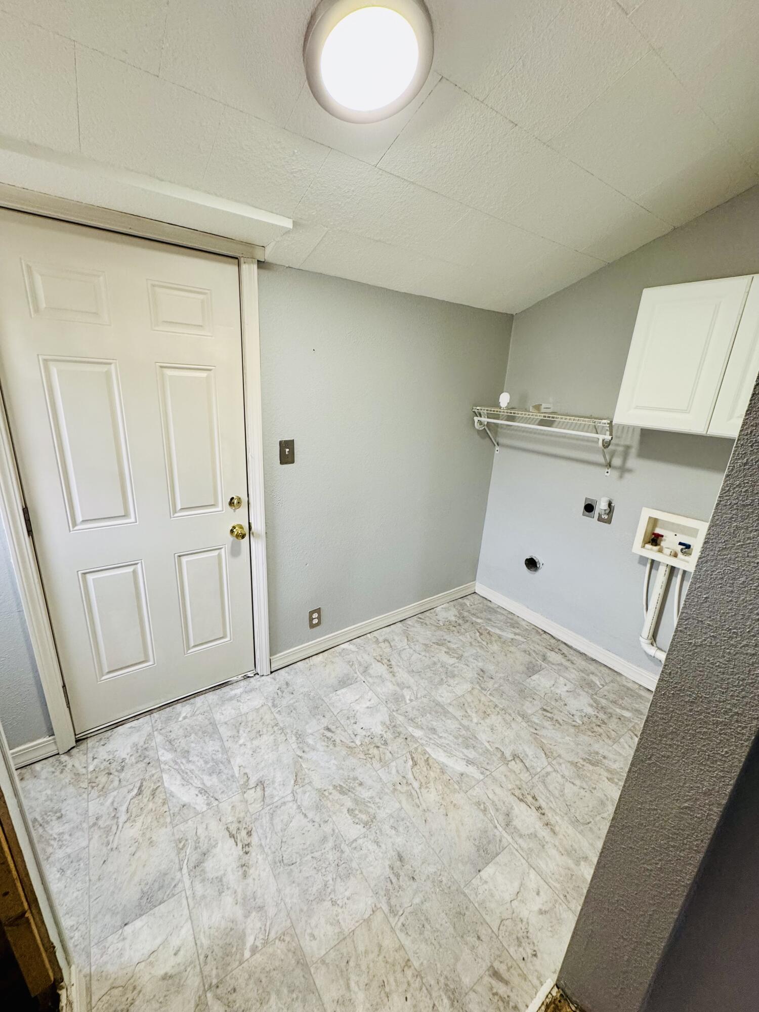 Empty laundry room with washer hookups, a wall shelf and cabinet, and marble-look tile flooring.