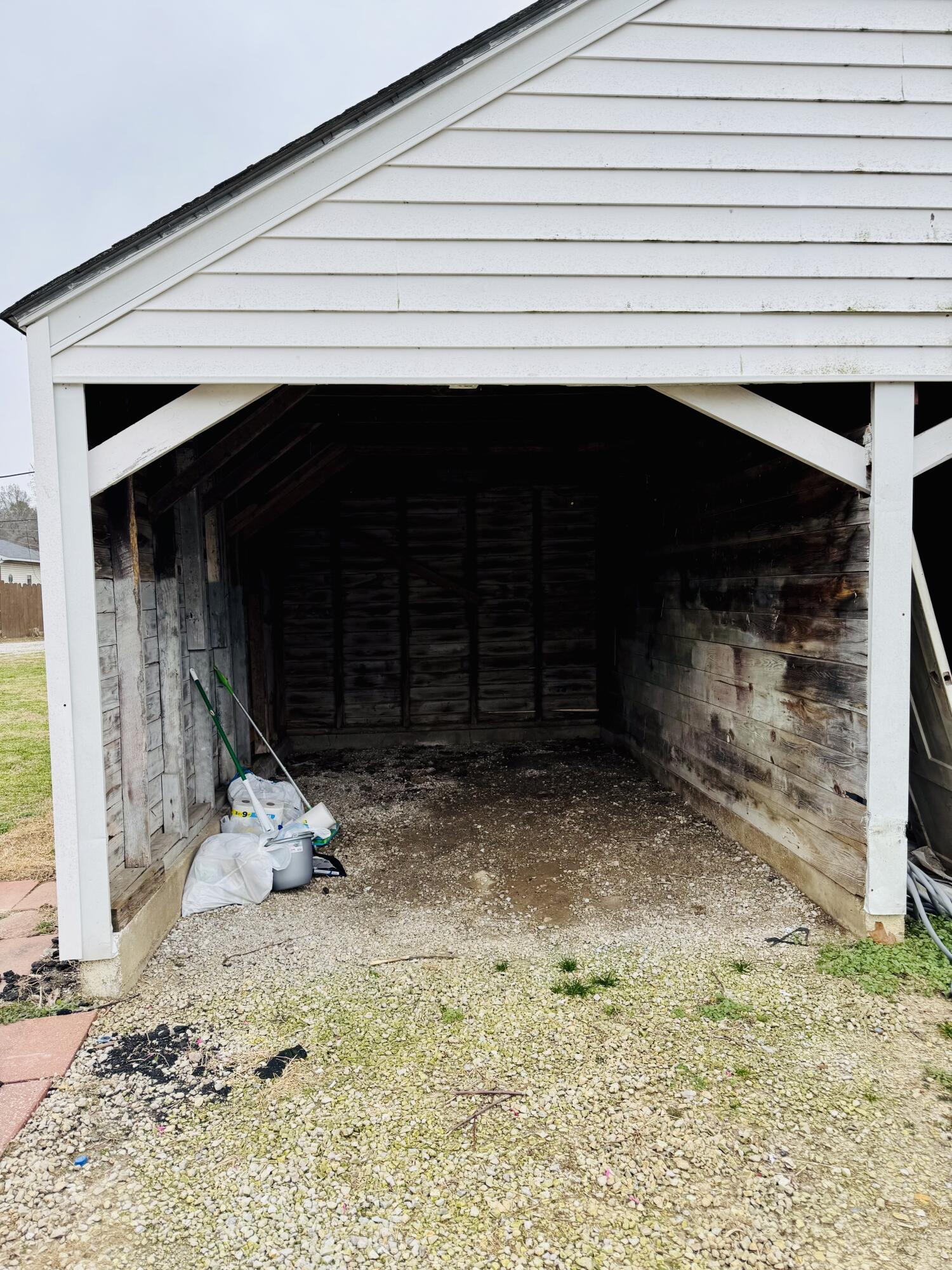 Open-sided wooden carport with a white gable roof, gravel floor, and cleaning supplies.