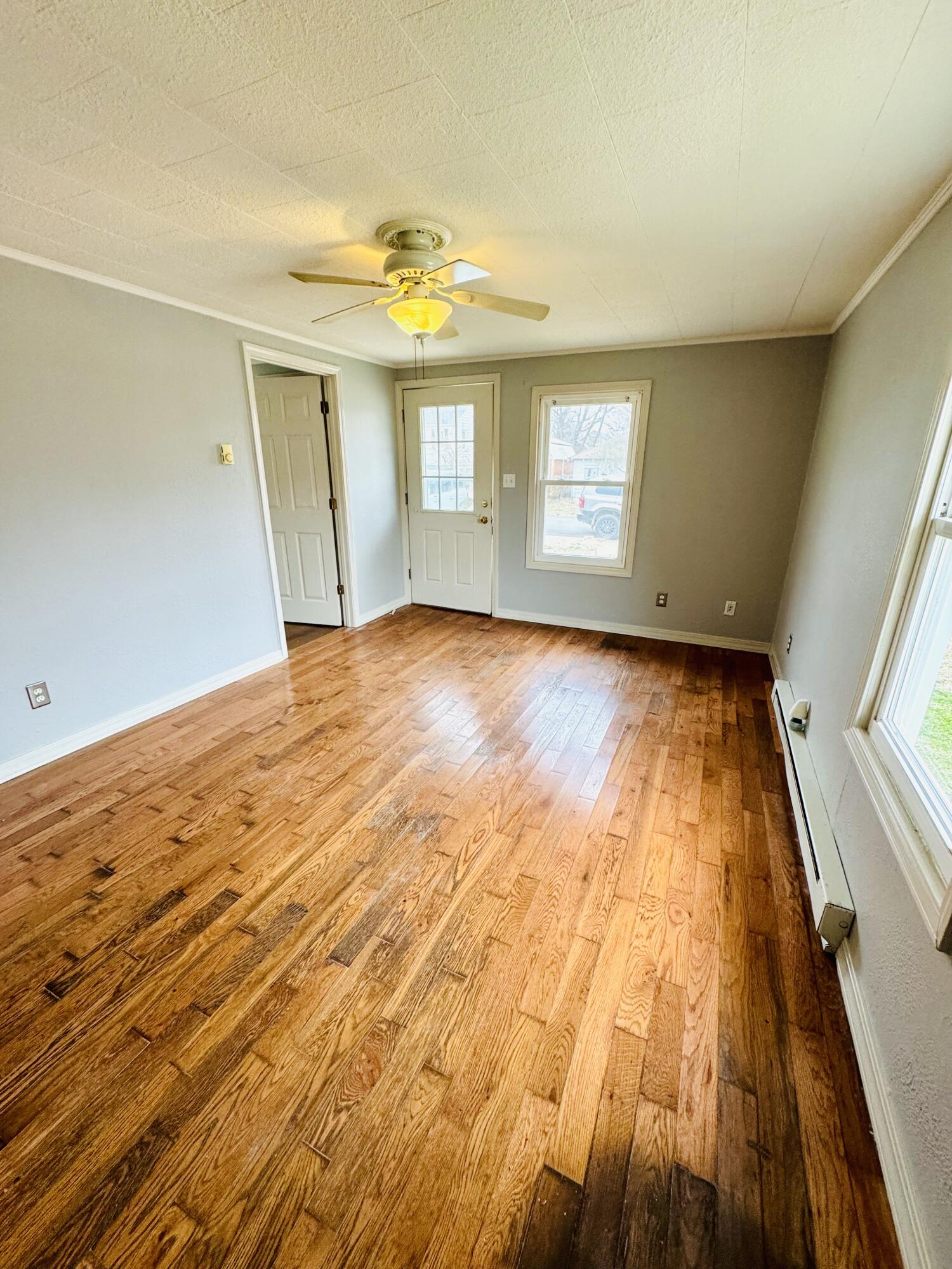 Empty room with warm hardwood floors, a ceiling fan, and a glass-paneled door beside a window.