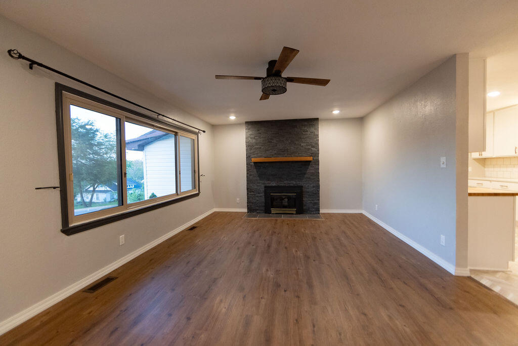 Empty living room with wood floors, a stone fireplace, and a large picture window.