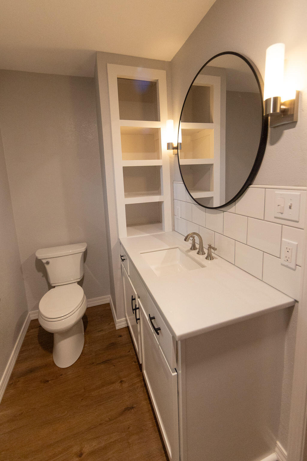 Bathroom with a white vanity, oval mirror, built-in shelves, and wood flooring.