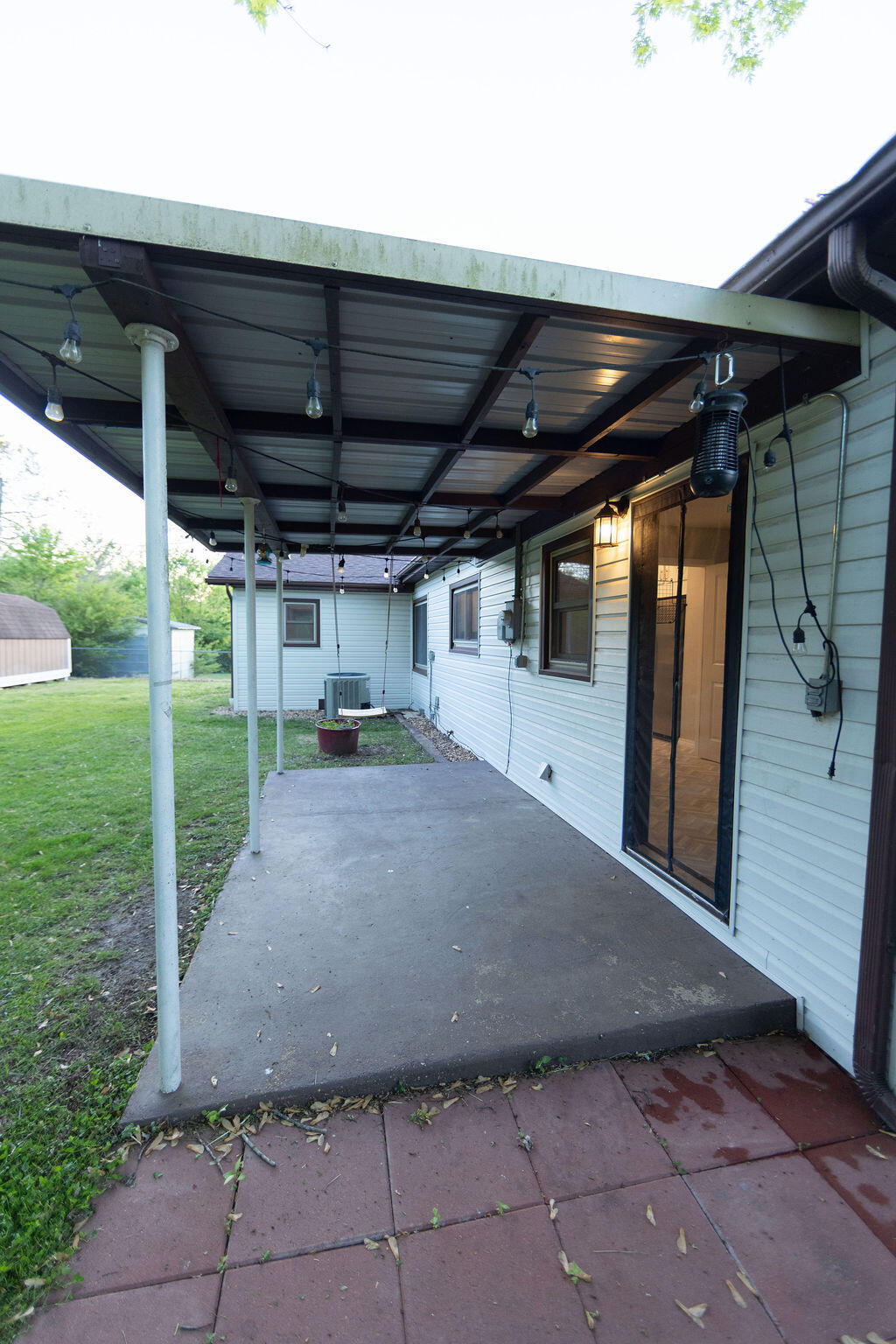 Covered concrete patio beside a light-sided house, with string lights and a sliding screen door.