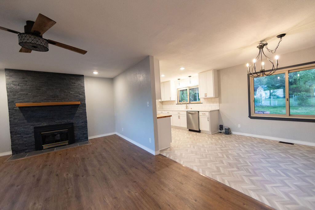 Open living and dining area with a stone fireplace, pale herringbone flooring, and an adjoining white kitchen.