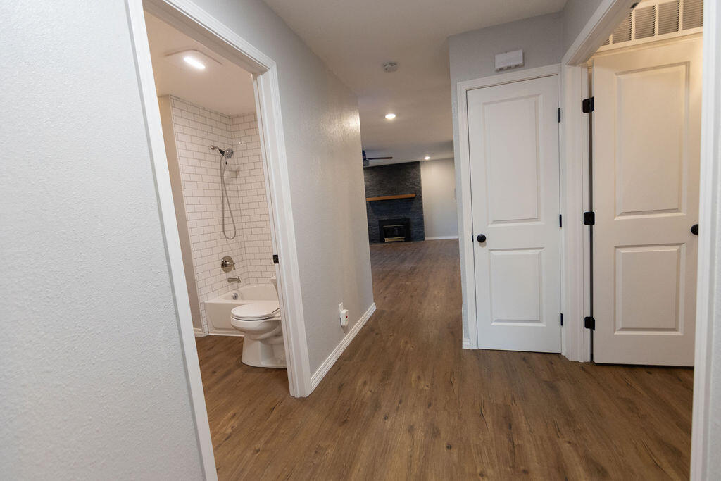 Empty hallway with wood-look floors, a tiled bathroom doorway, and a stone fireplace in the distance.