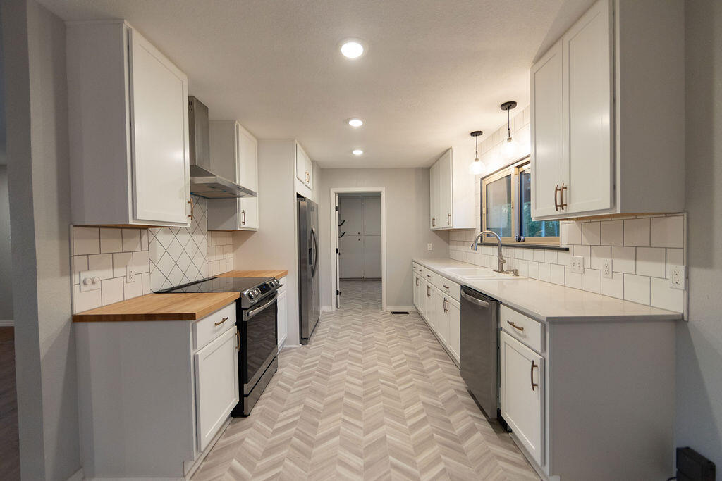 Long white kitchen with stainless steel appliances, butcher-block counter, and herringbone-pattern flooring.