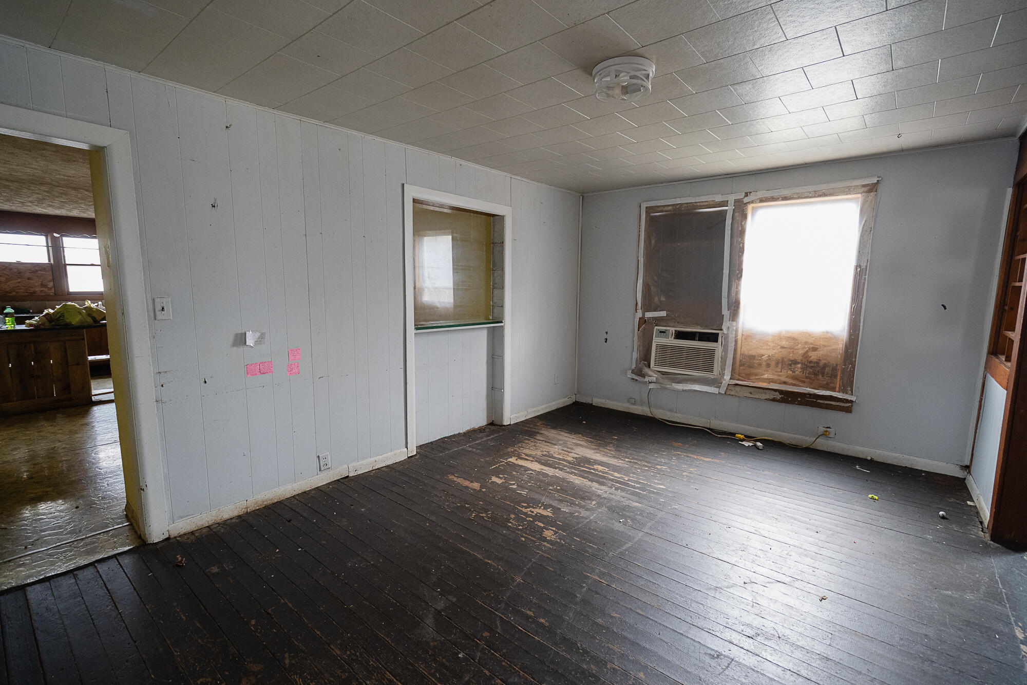 Vacant room with worn plank flooring, paneled walls, a pass-through opening, and a window air conditioner.