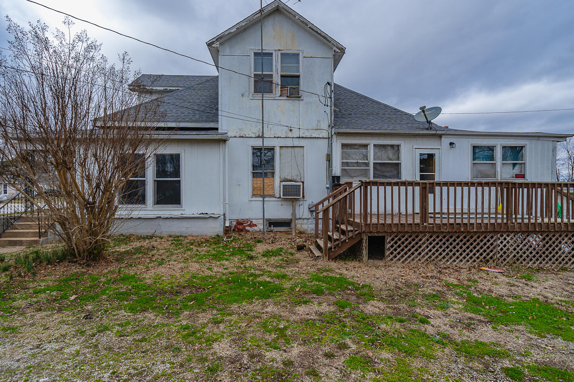 Rear of a weathered white house with a raised wooden deck and leafless tree.