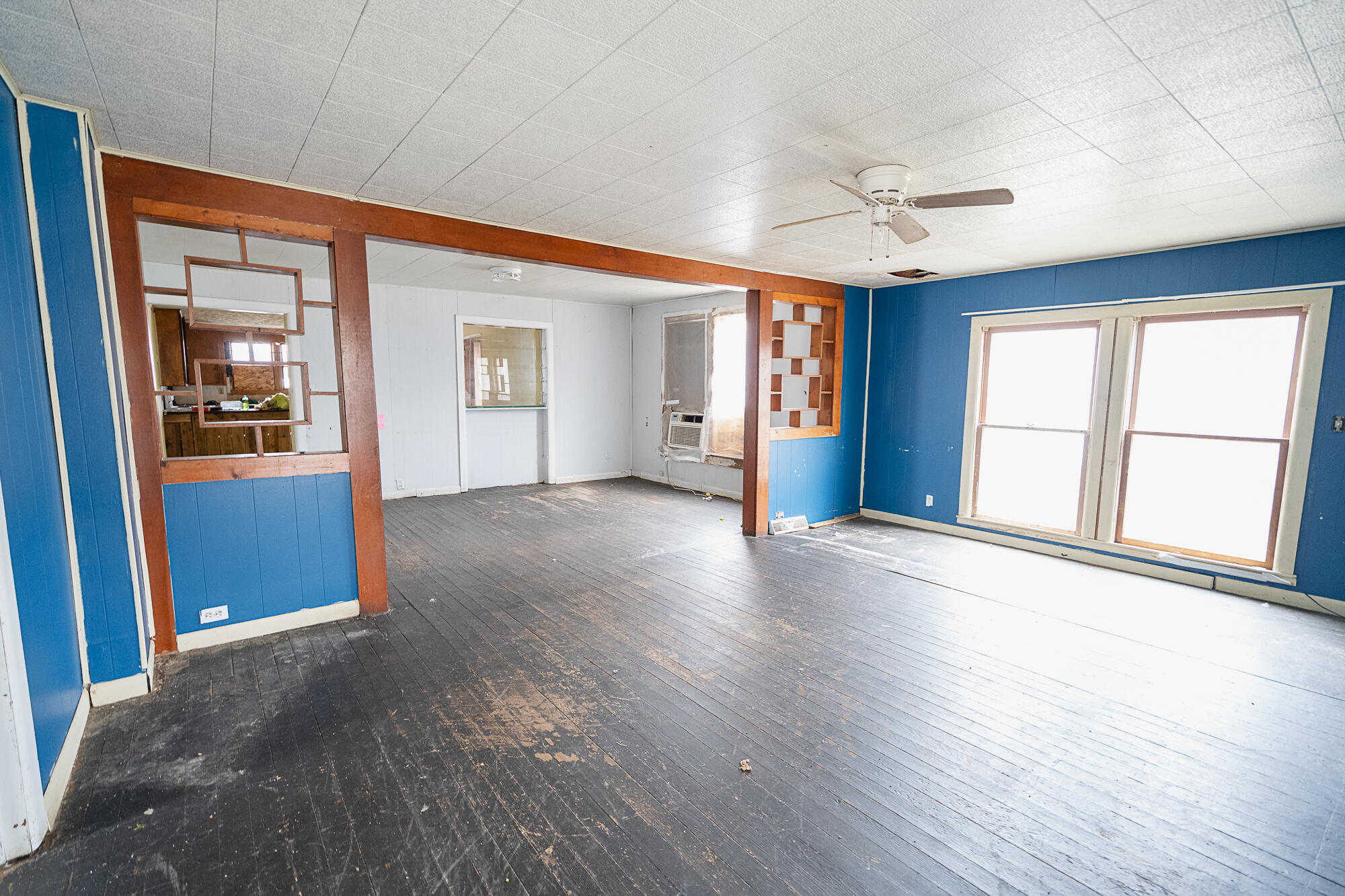 Empty living room with blue walls, worn wood floors, and large paired windows.
