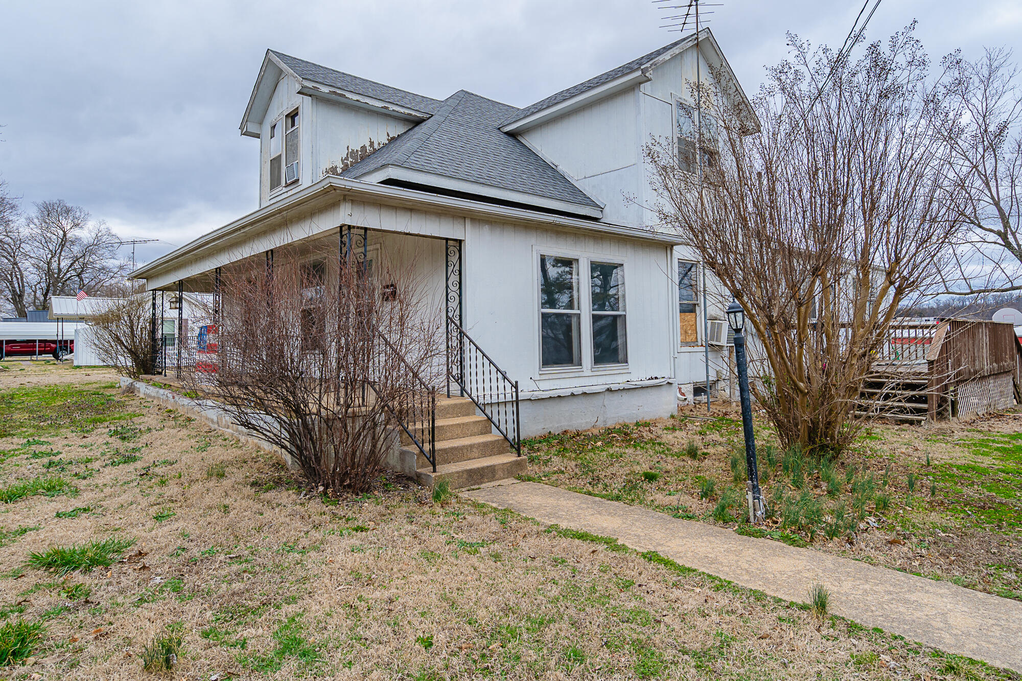 White two-story house with a covered porch, peeling paint, and leafless shrubs.