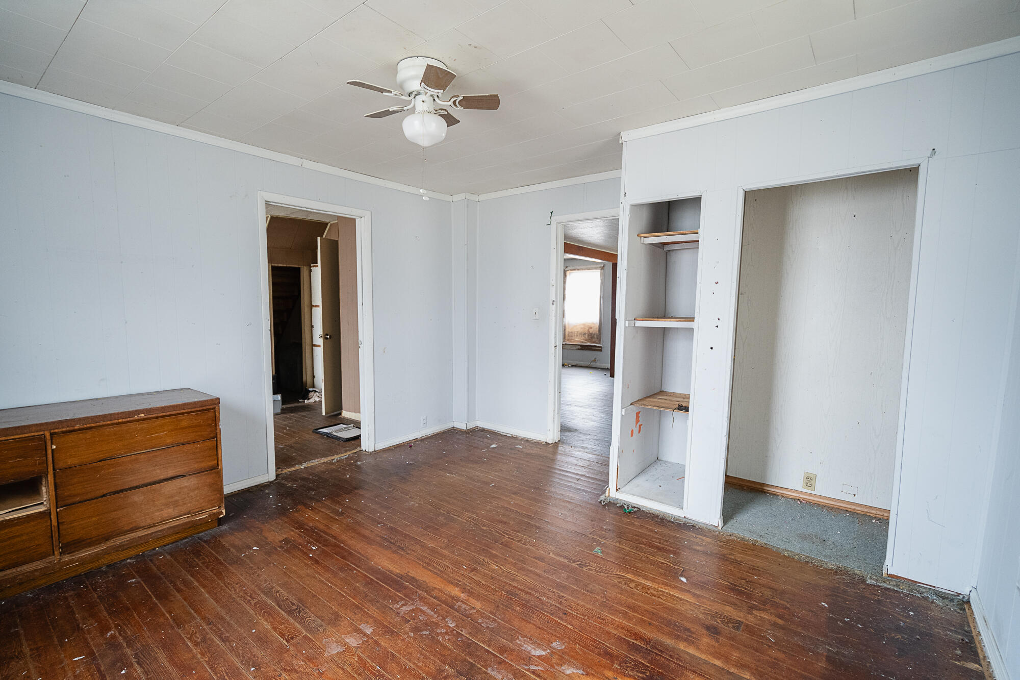 Vacant room with worn wood floors, built-in closet shelving, and a ceiling fan.