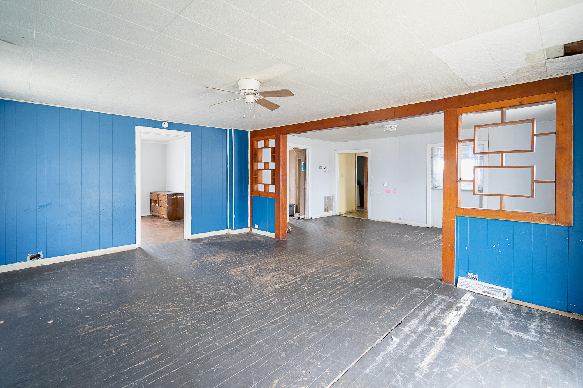 Vacant living room with blue paneled walls, worn dark flooring, and a wooden geometric room divider.
