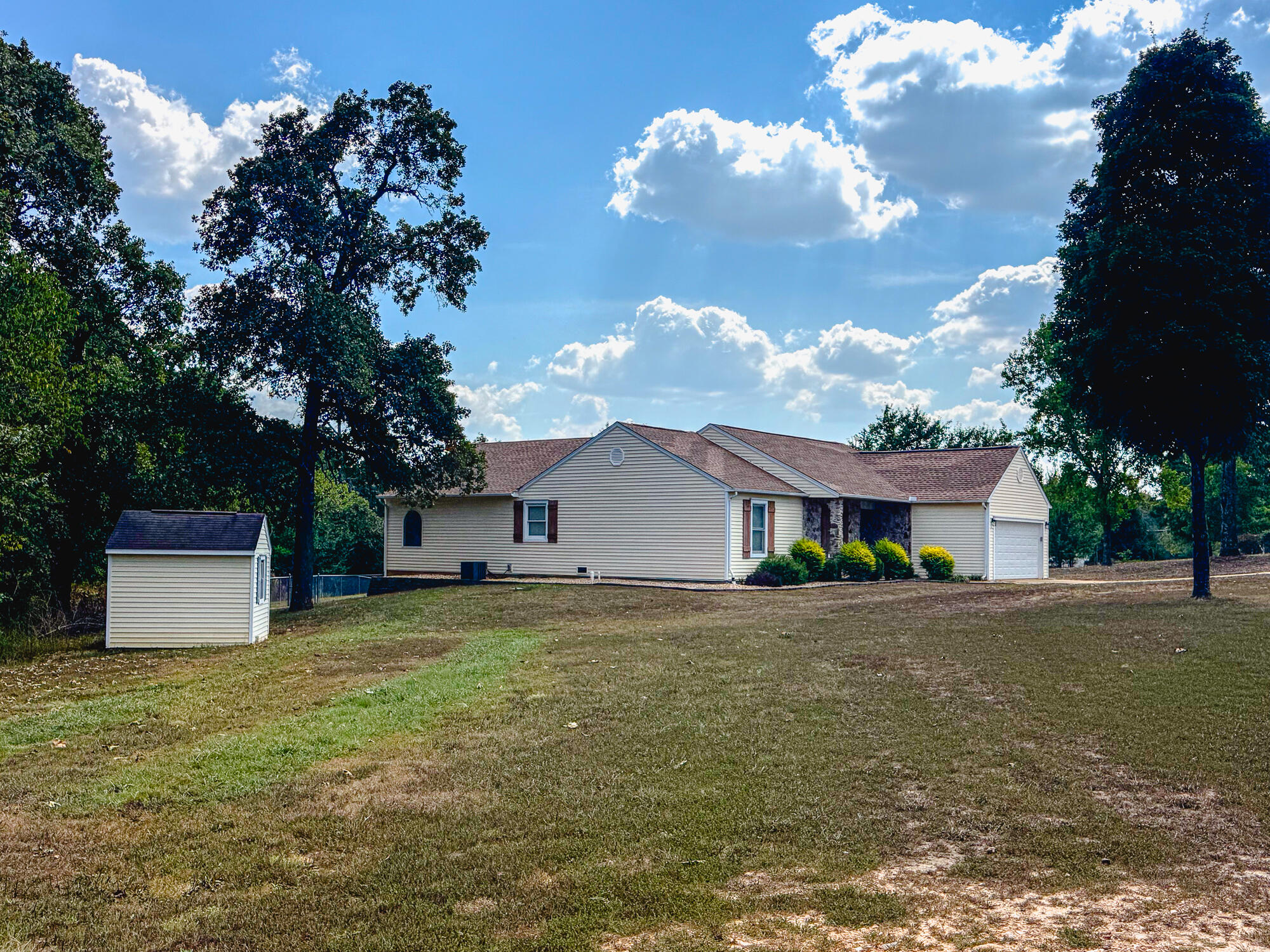 Single-story home with an attached garage and small matching shed beside a broad lawn and mature trees.