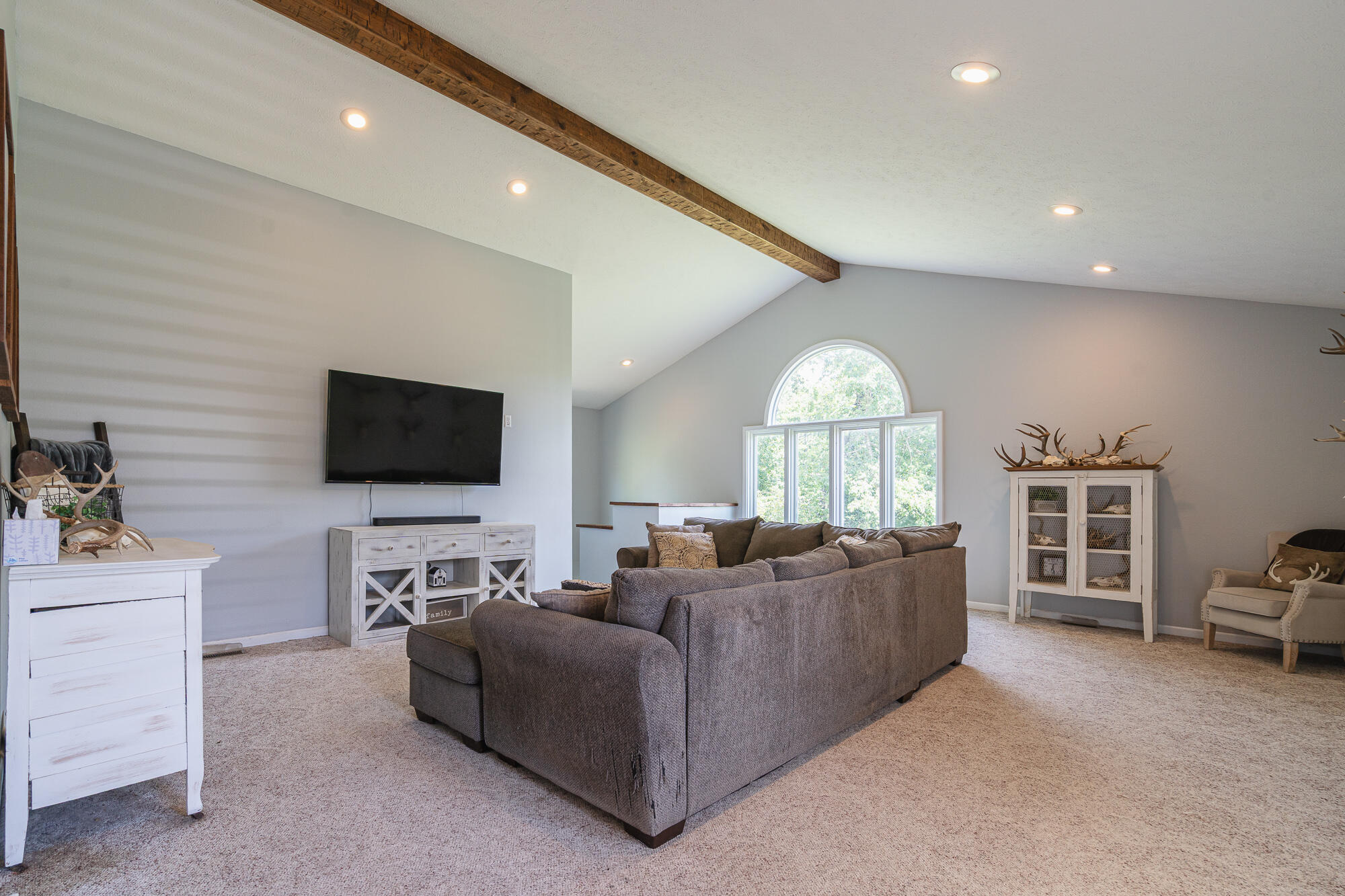 Furnished living room with a vaulted ceiling, exposed wood beam, gray sectional, and arched window.