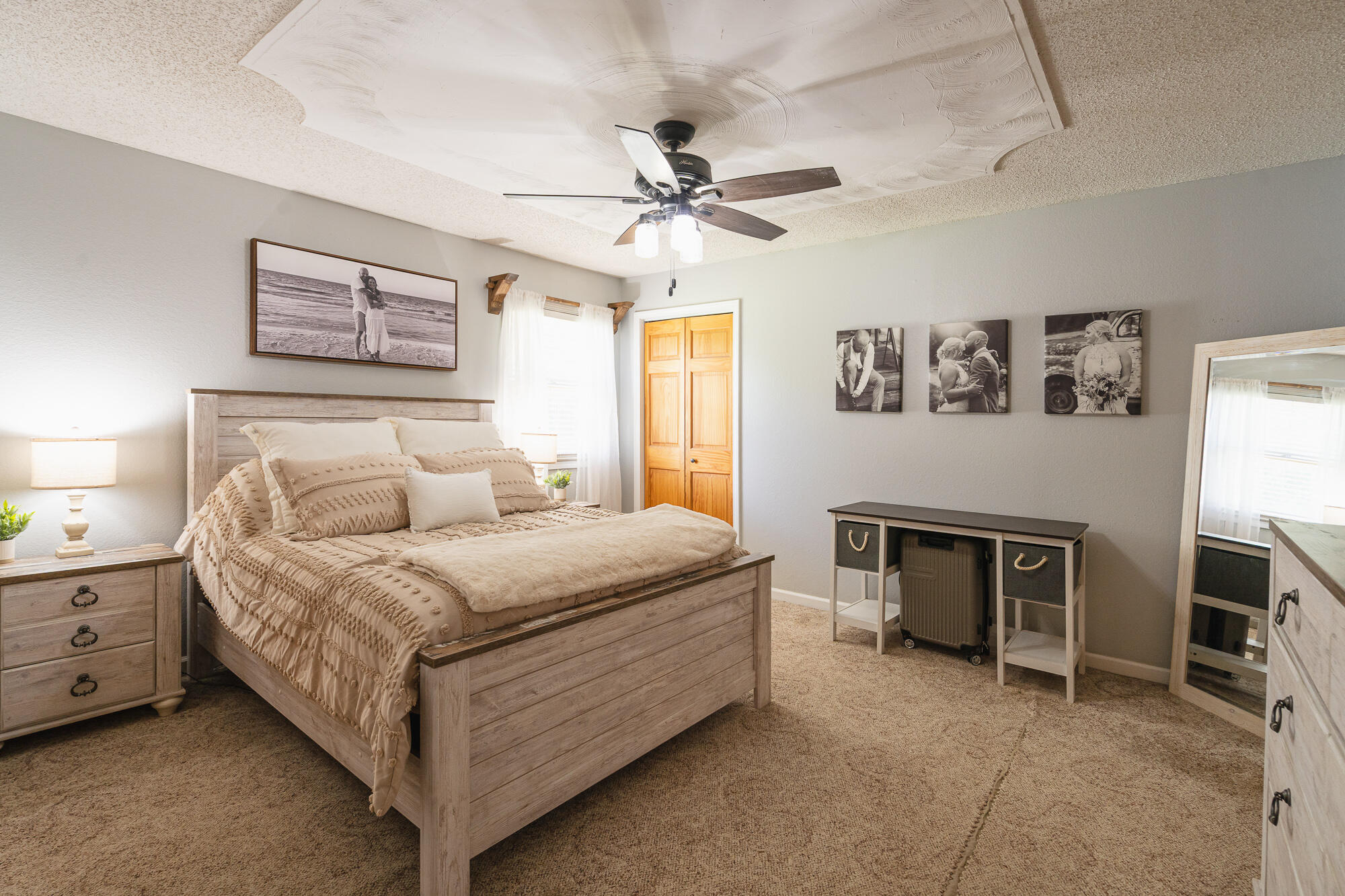 Furnished bedroom with a light wood bed, beige bedding, ceiling fan, and framed photographs.