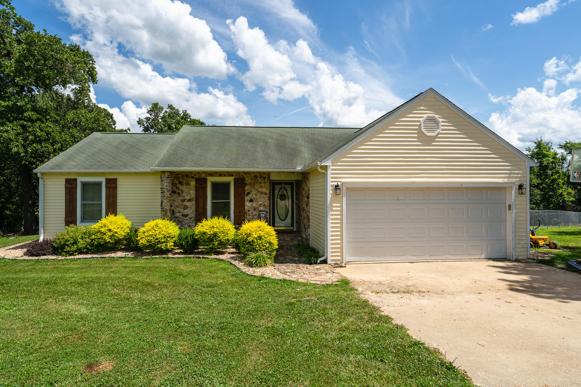 Beige single-story house with a stone entry, two-car garage, and bright yellow shrubs along the front.