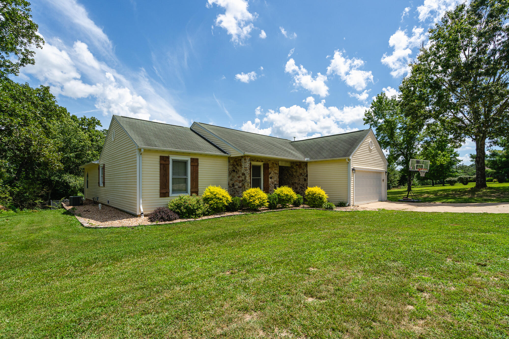 Cream-sided home with a stone entry, attached garage, and basketball hoop beside the lawn.