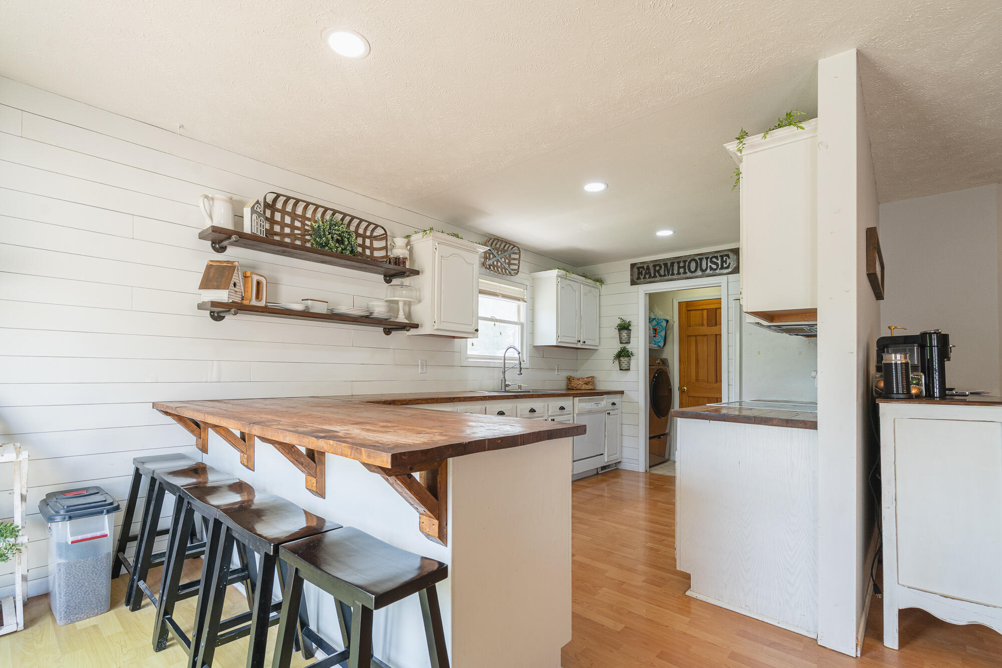 Farmhouse-style kitchen with a large wood-topped island, dark stools, white shiplap walls, and open shelves.