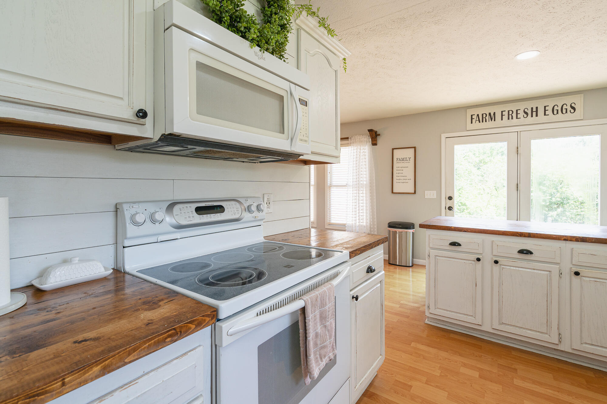 Kitchen with white cabinetry, dark wood counters, an electric range, and a glass door beneath a “Farm Fresh Eggs” sign.