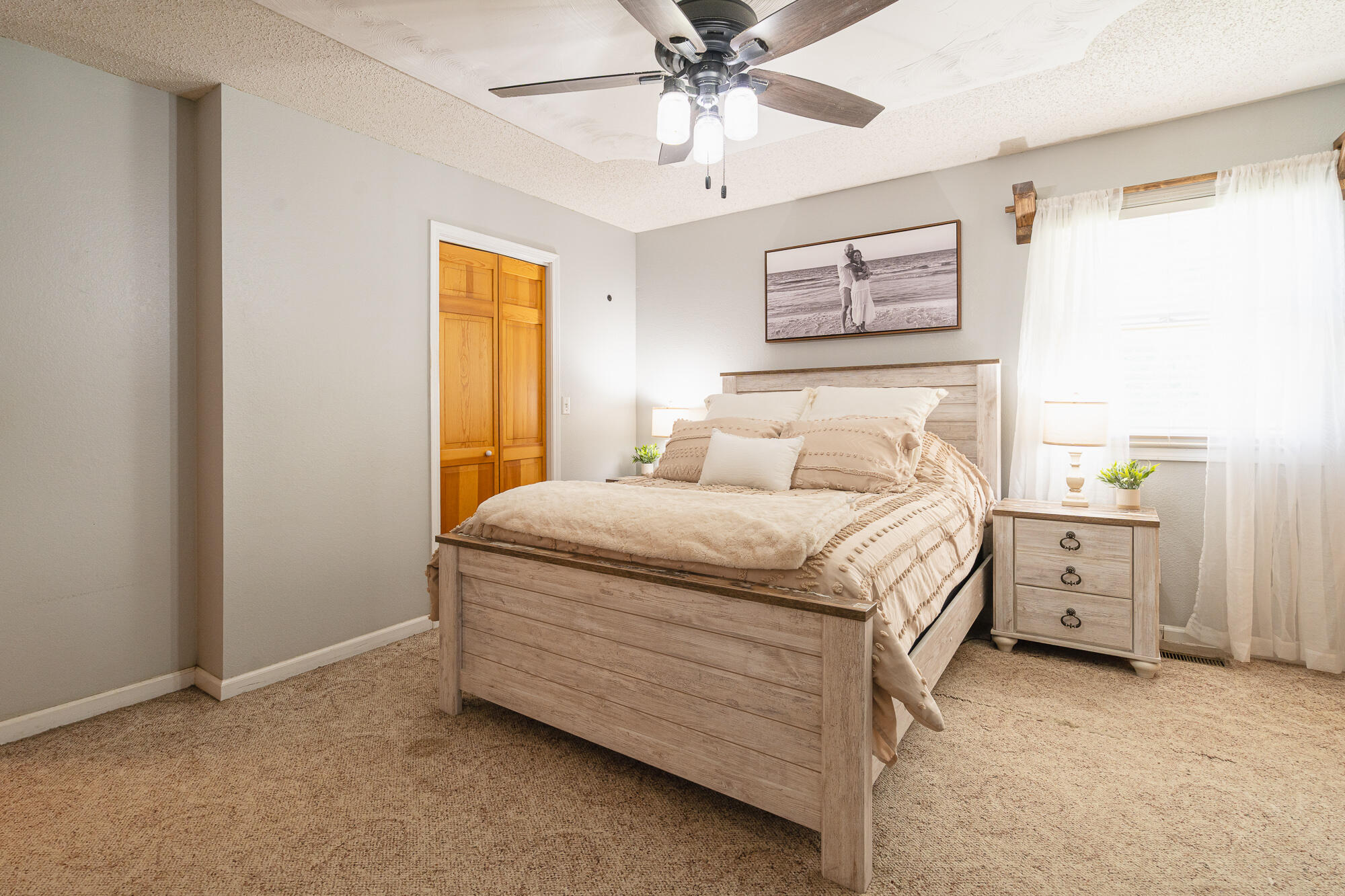 Bedroom with a light wood bed, beige bedding, sheer curtains, and a ceiling fan.