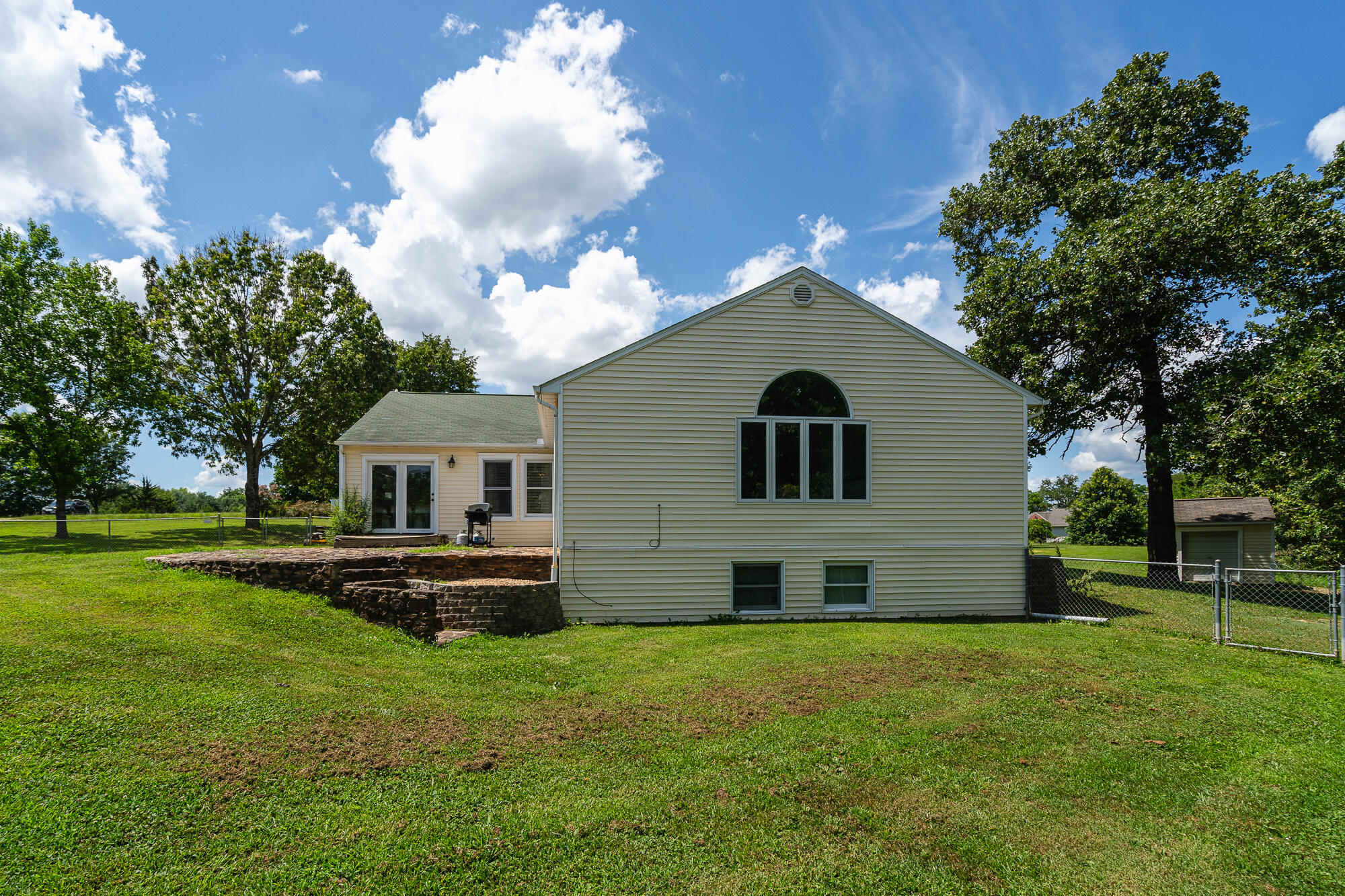 Rear of a light-sided house with an arched window, stone patio wall, broad lawn, and mature trees.