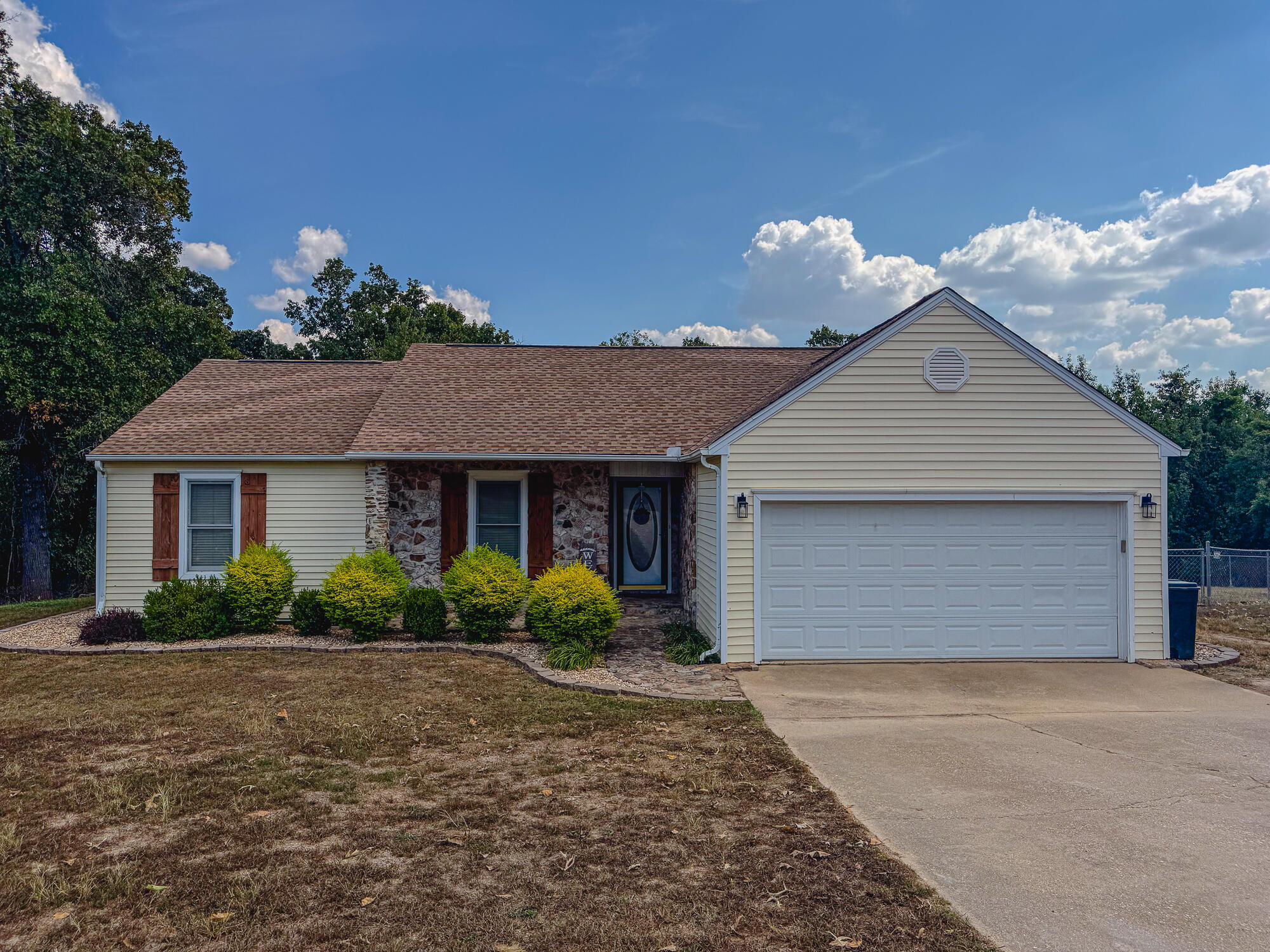 Single-story beige home with stone entry, red-brown shutters, and an attached two-car garage.