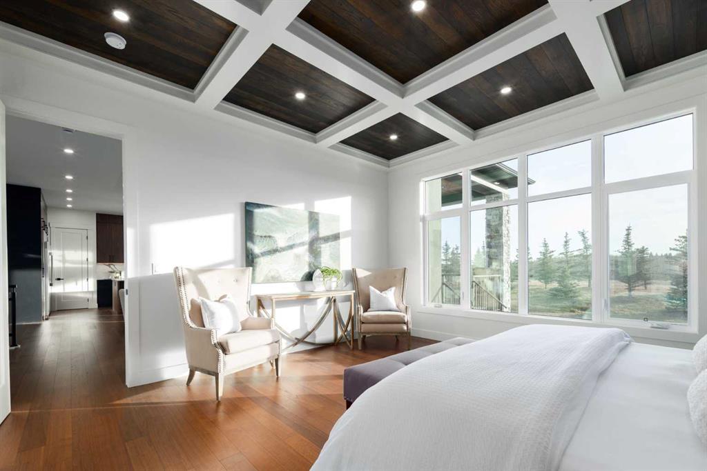 Bedroom with a sitting area, dark wood coffered ceiling, and large windows overlooking trees.
