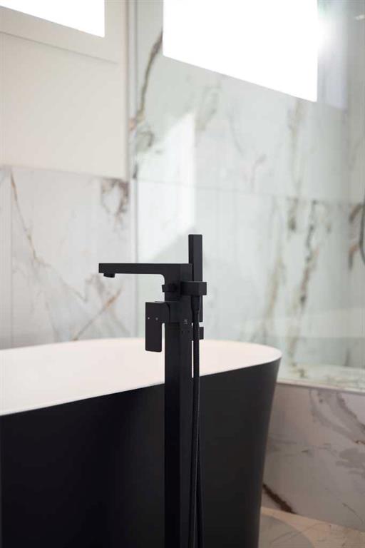 Freestanding bathtub with a matte-black faucet against marble-look tile and tall windows.