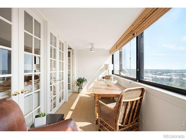 Furnished sunroom with French doors, a wooden table, wicker chairs, and expansive city-facing windows.