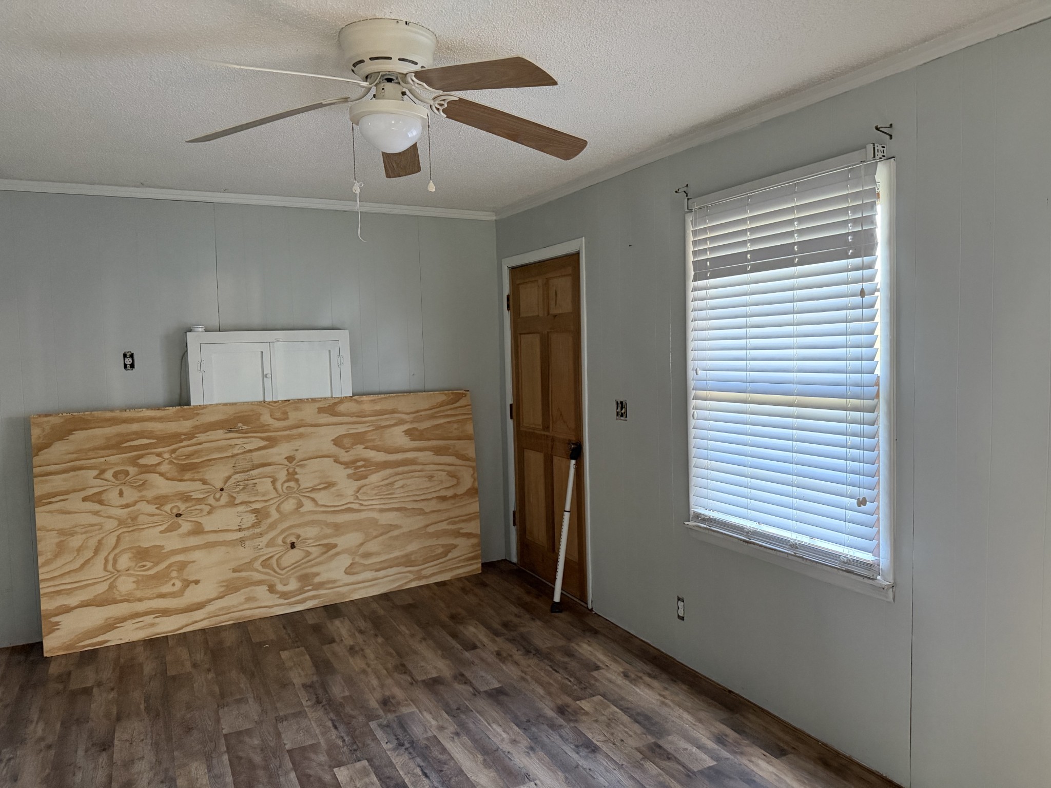 Empty room with gray paneled walls, dark flooring, a ceiling fan, and plywood leaning against the wall.