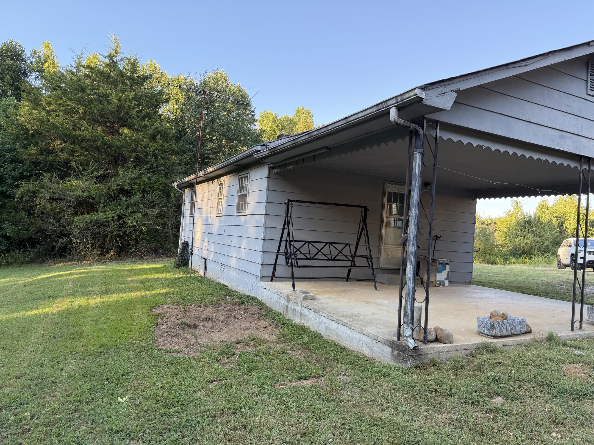Gray-sided house with a covered porch, metal swing, and grassy yard beside dense trees.