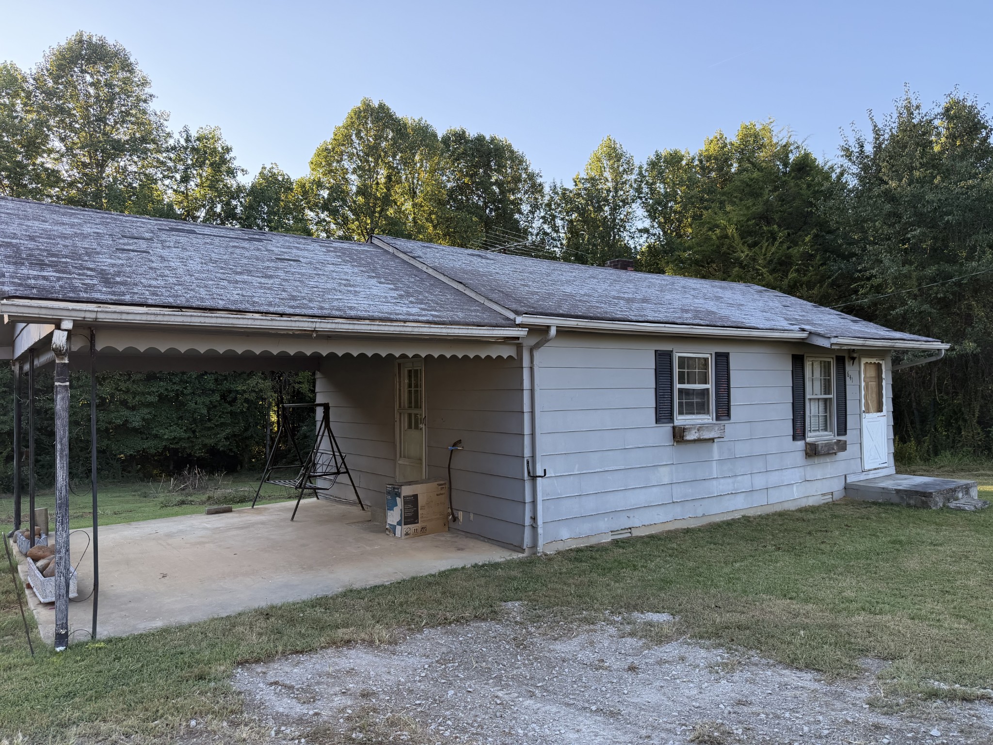 Single-story gray house with a broad attached carport and wooded surroundings.