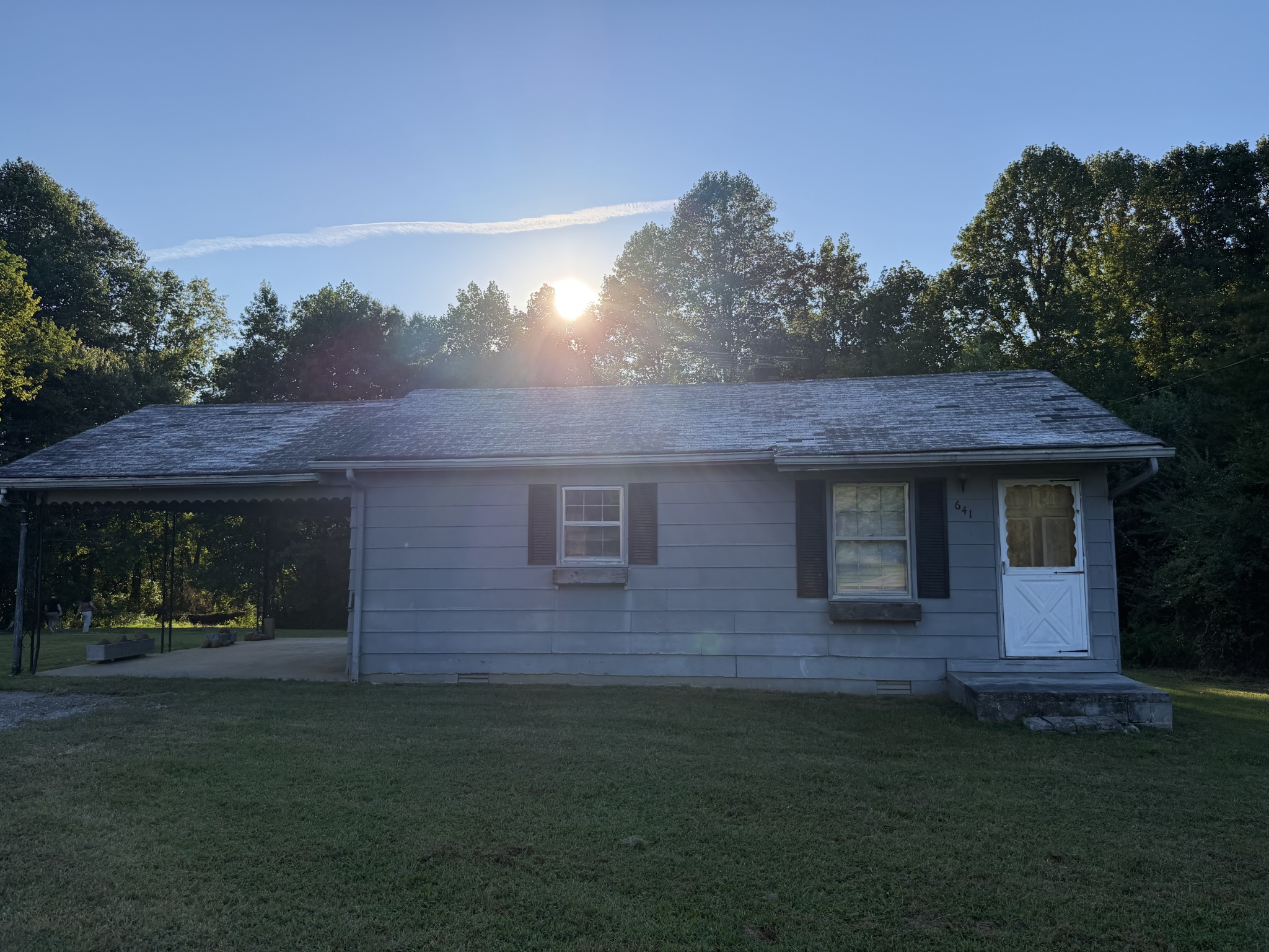 Single-story gray house with an attached carport, photographed at golden hour among trees.