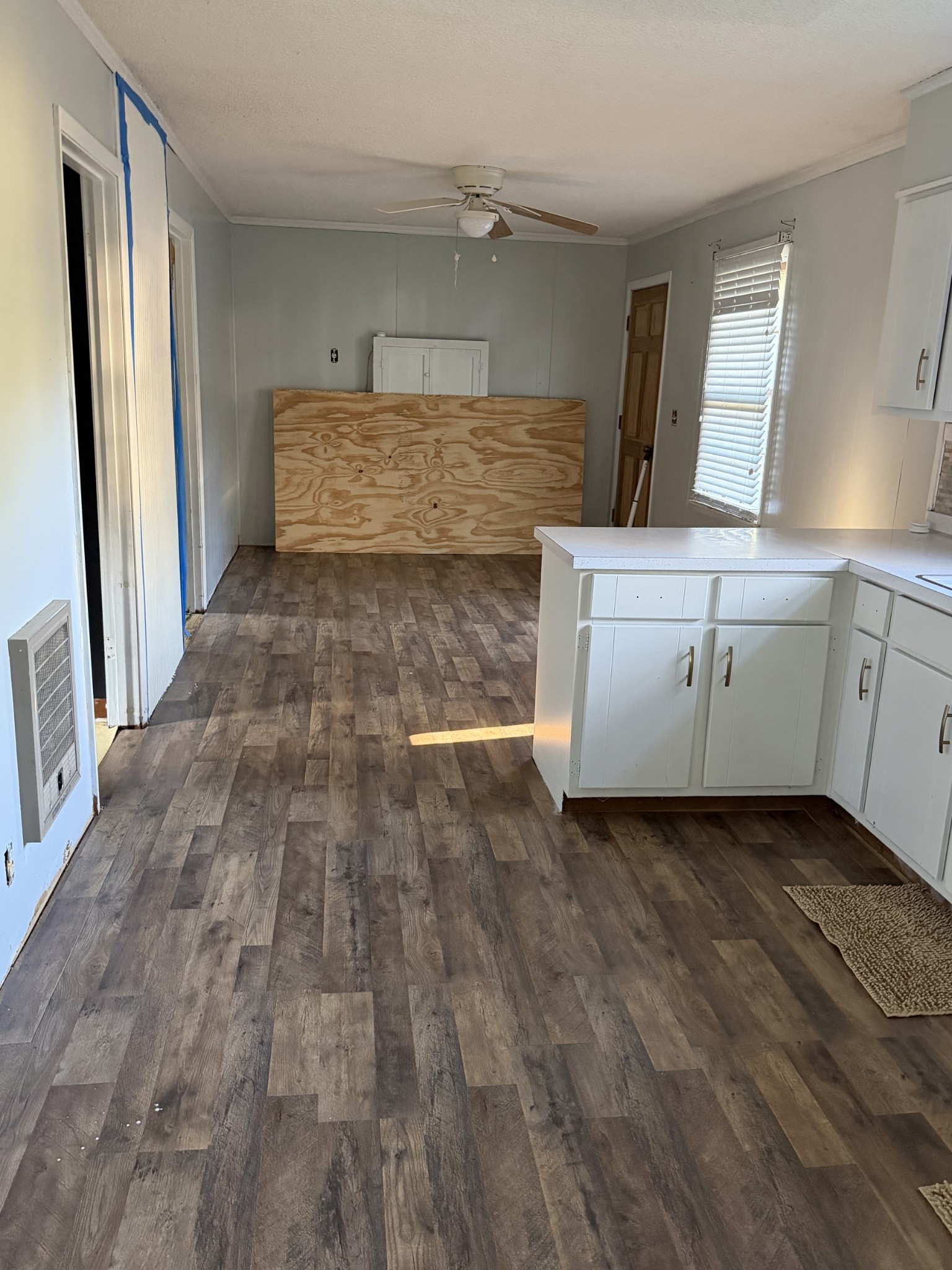 Kitchen and adjoining room with white cabinets, dark wood-look flooring, and plywood covering the back wall.
